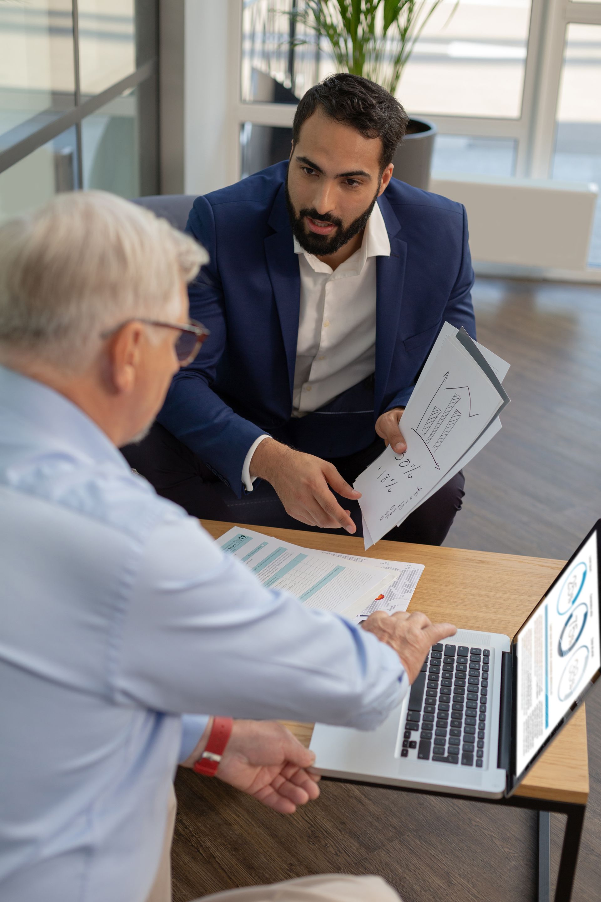 Man in a suit points at document for older man, who uses a laptop at a table. Light-filled office setting.