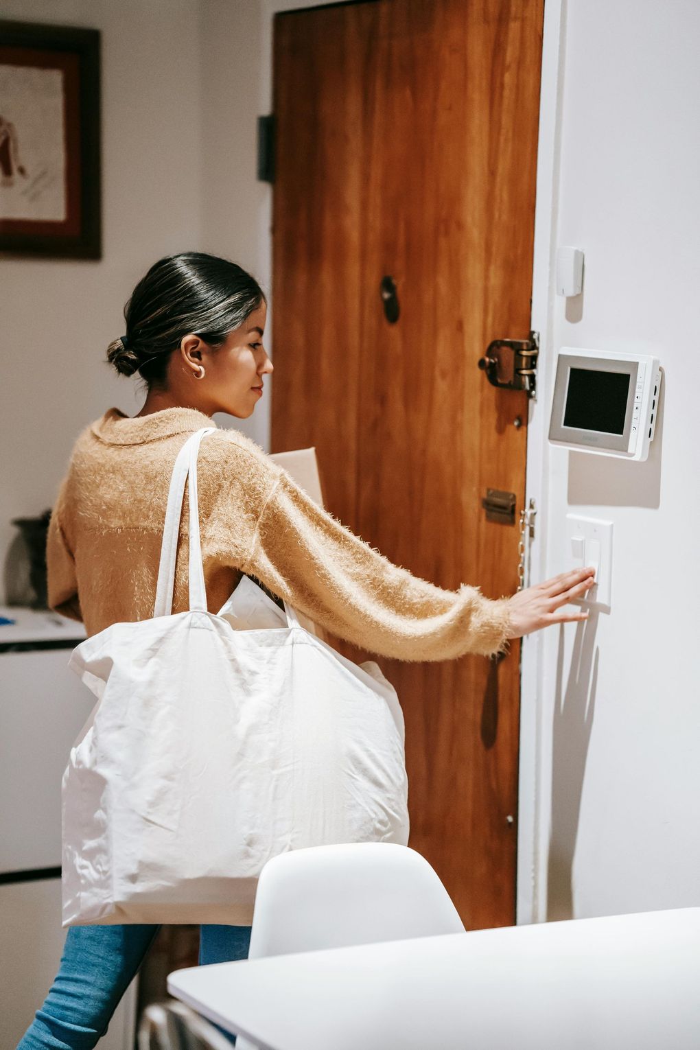 Woman entering a home, holding a tote bag, reaching for a light switch by a wooden door.