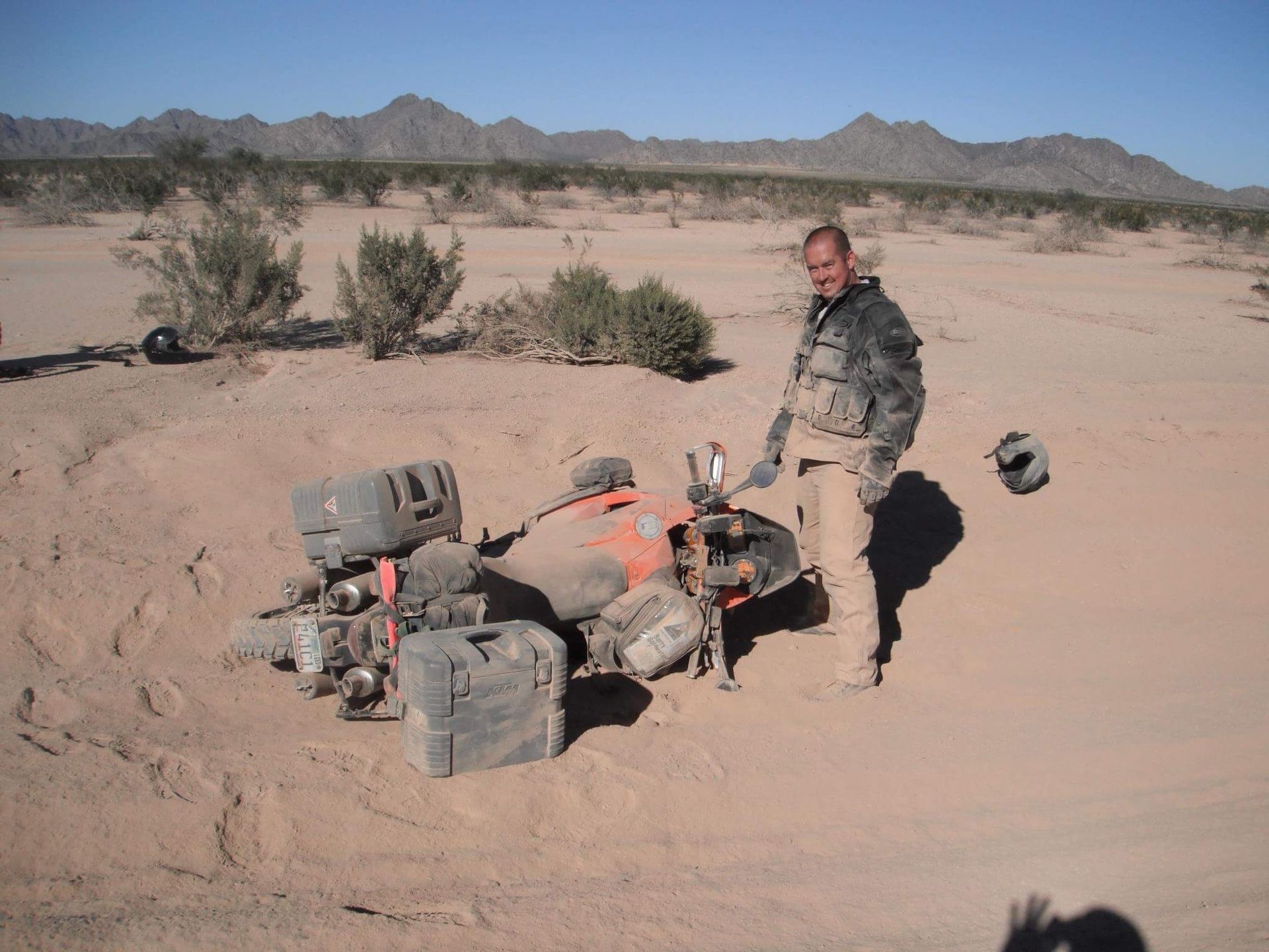 Man standing next to a bike laying on it's side in the dirt/desert. Man is dusty. 
