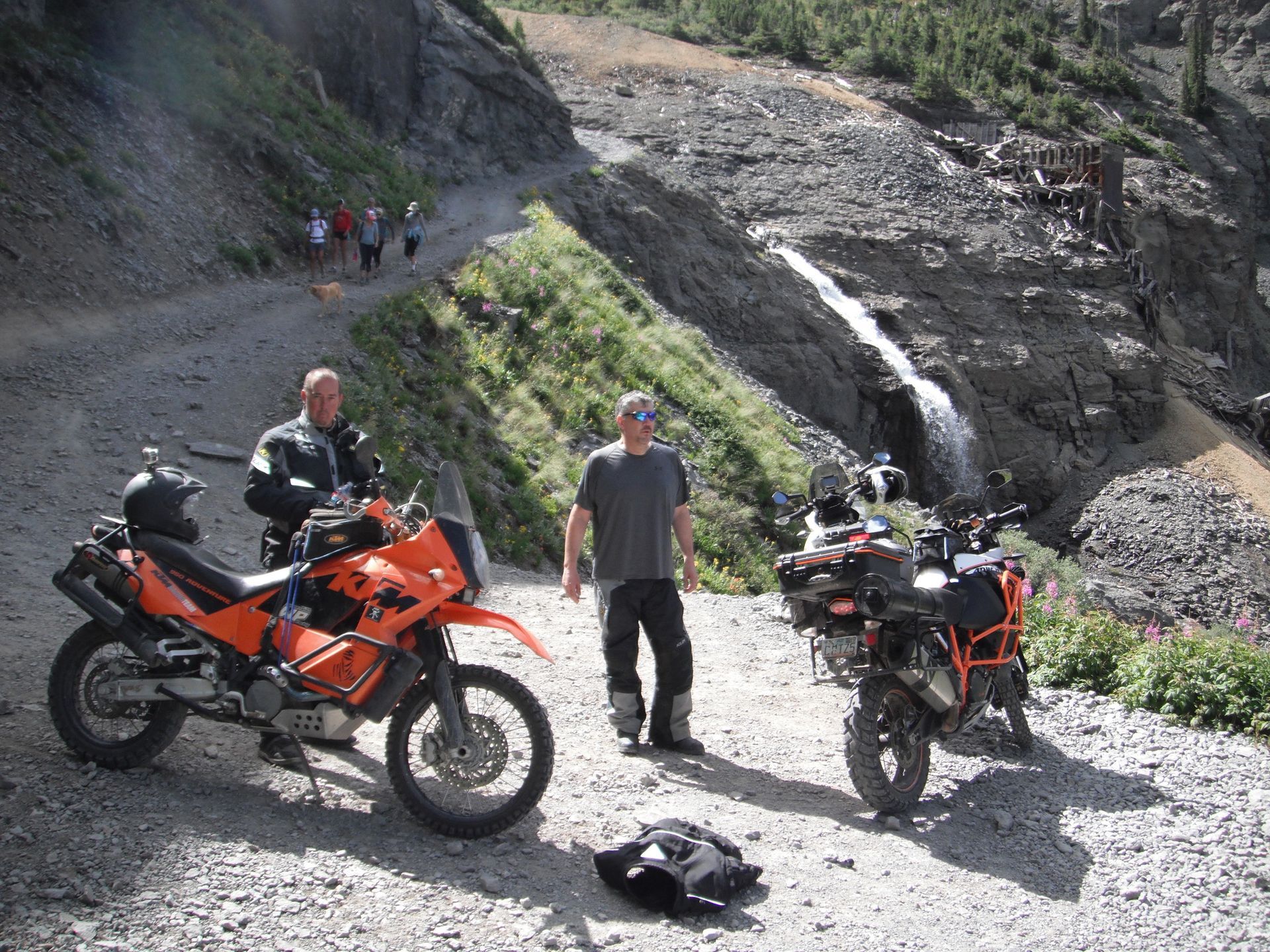 Two men are standing next to motorcycles on a dirt road near a waterfall.