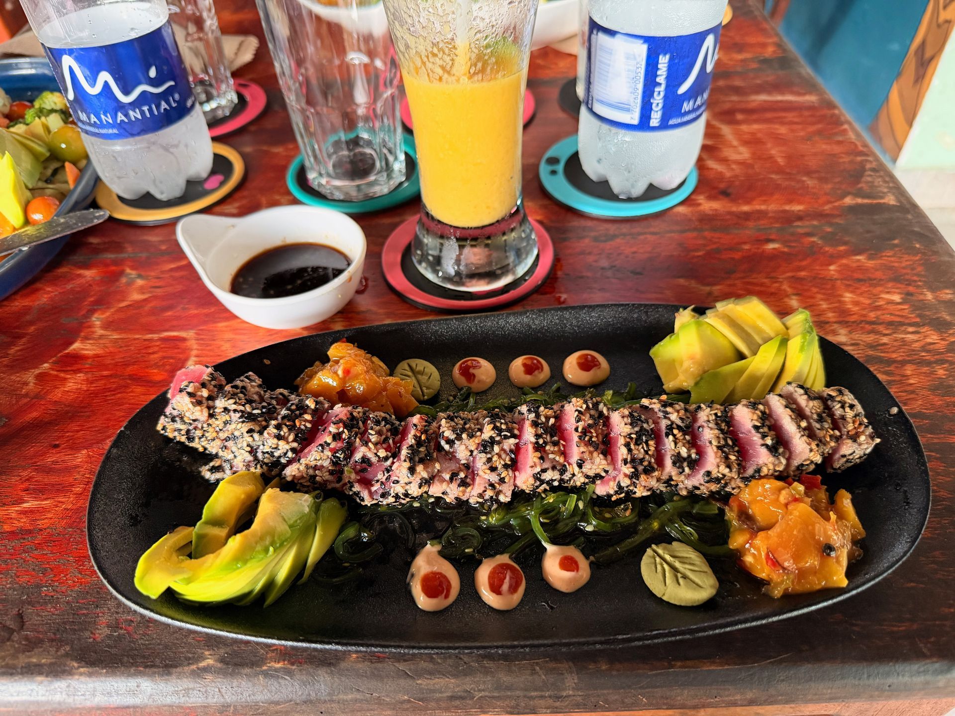 Seared tuna with sesame seeds, avocado, and sauce on a black plate, table setting with drinks.