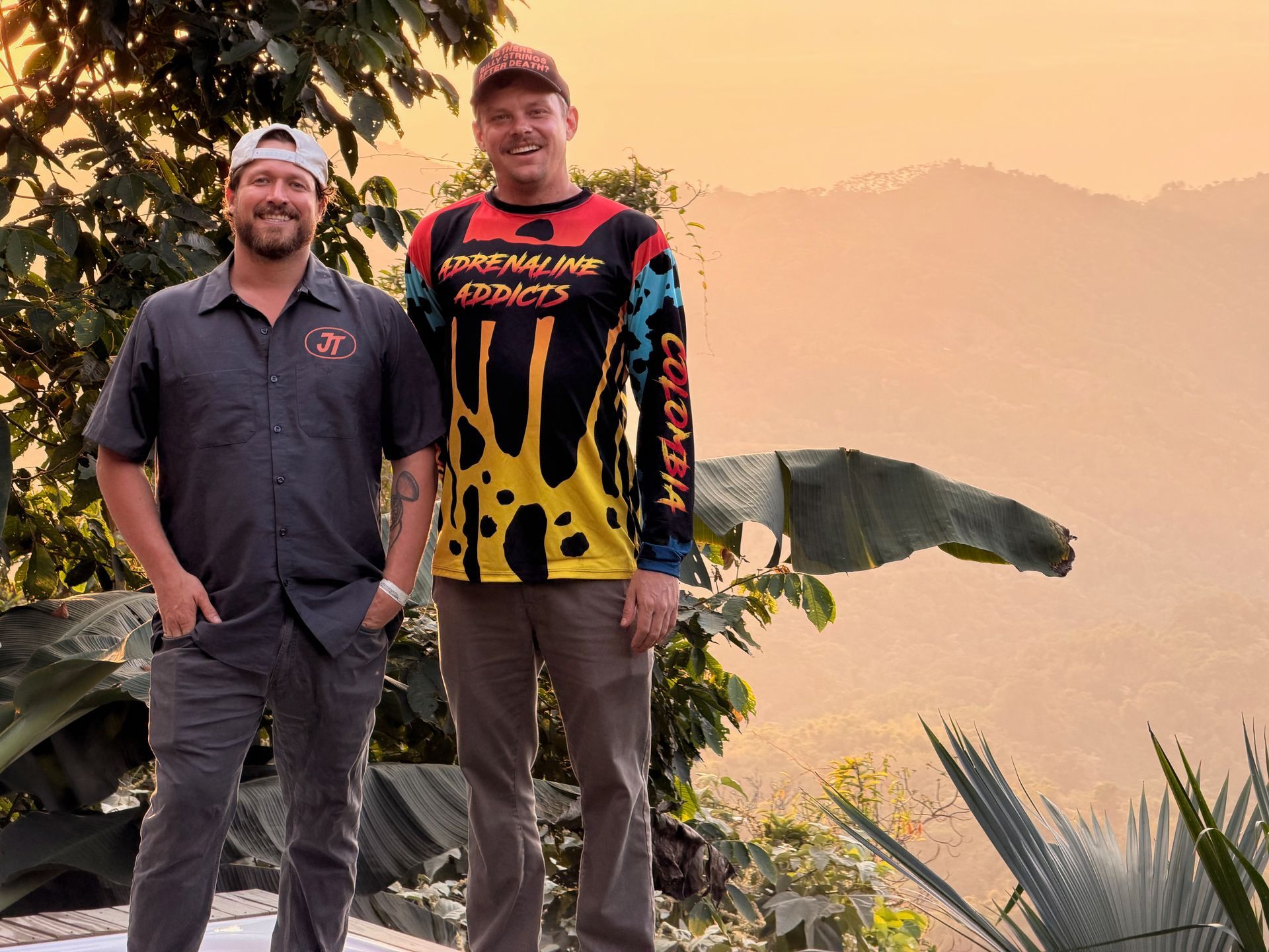 Two men standing outdoors with a sunset backdrop. One in a gray shirt, the other in a colorful jersey and khaki pants.