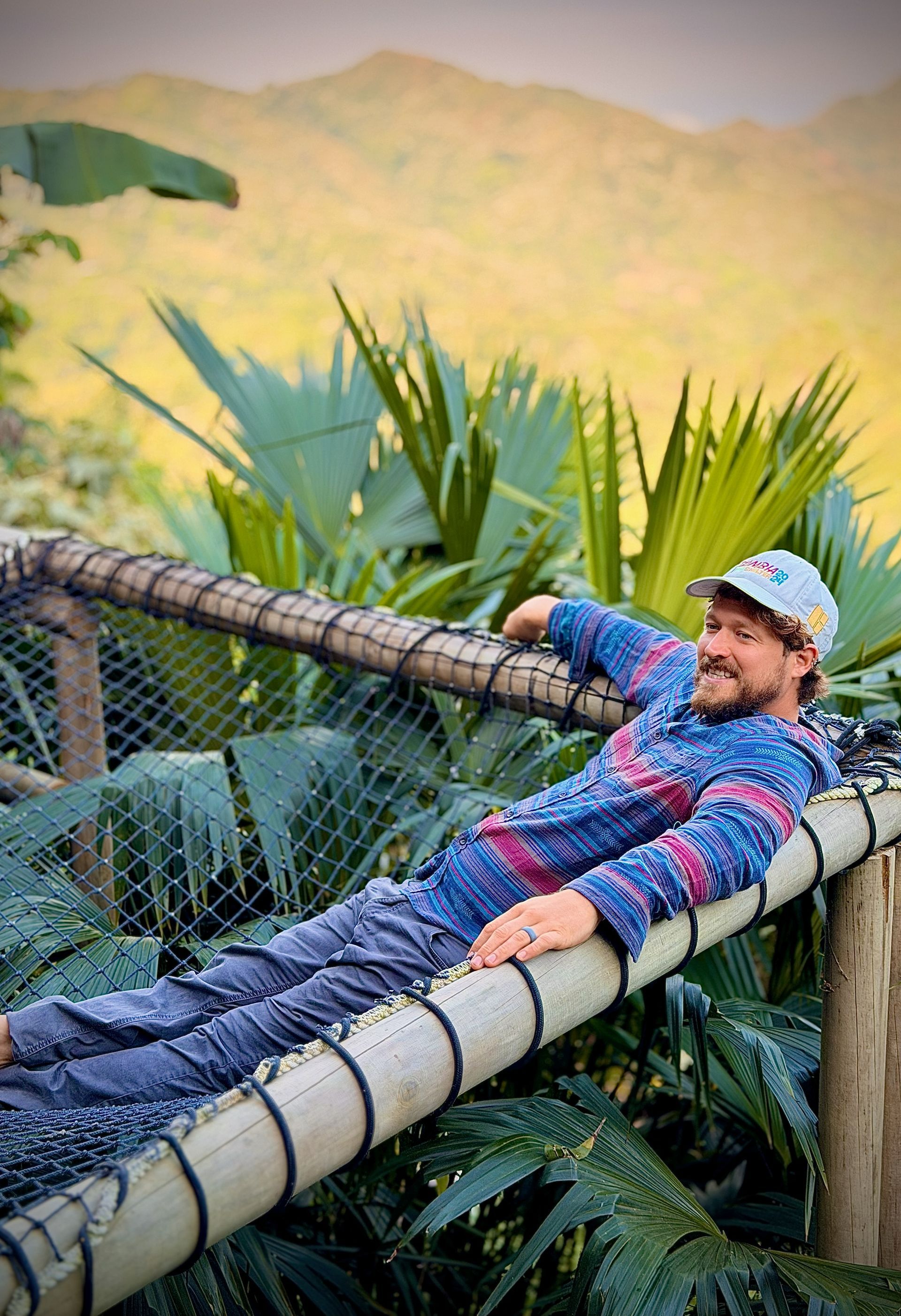 Man smiling, relaxing on a hammock-like structure, jungle background.
