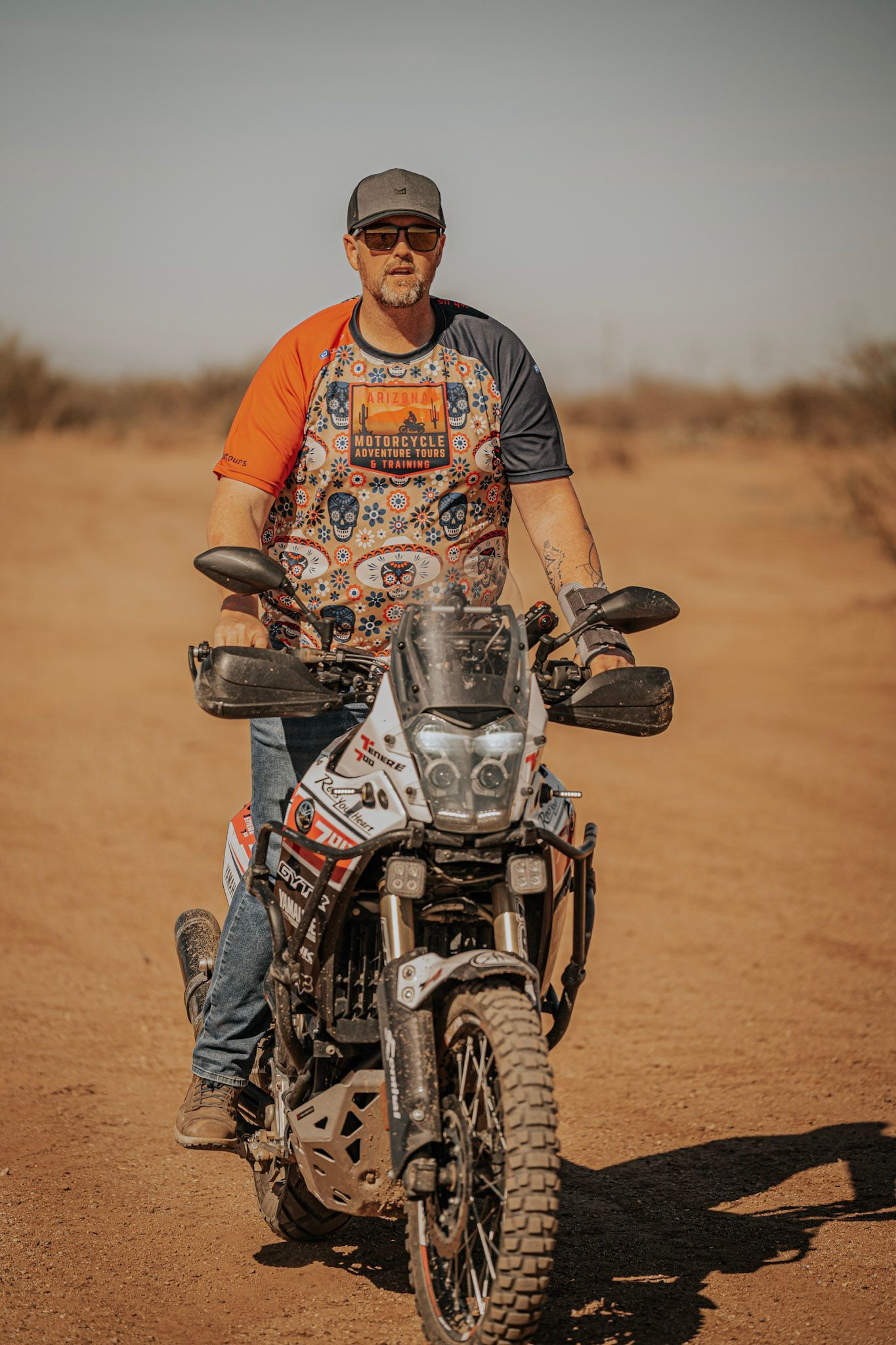 Man on a motorcycle in a desert setting; wearing a hat and colorful shirt; bike has off-road tires.