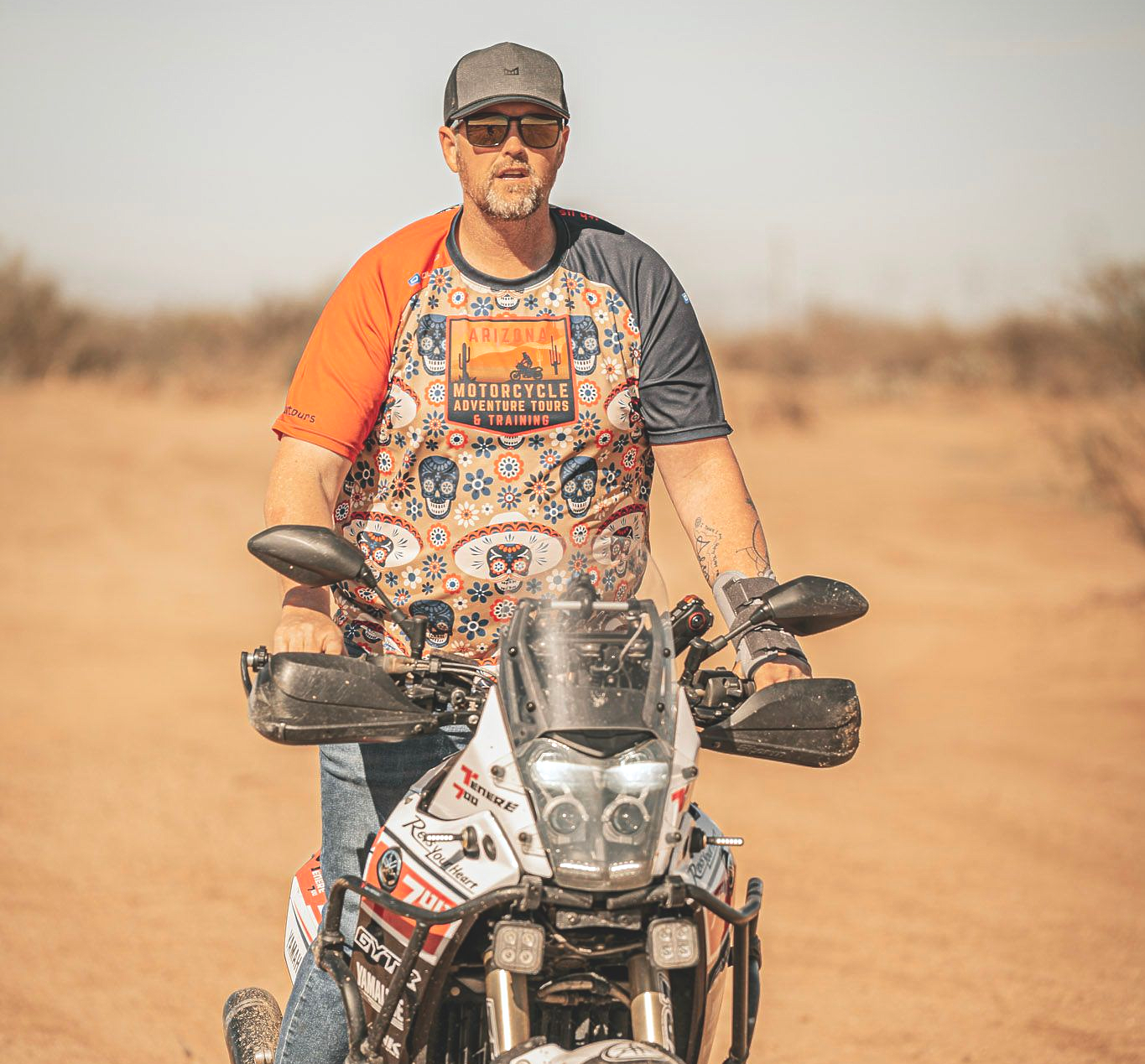 Man on a motorcycle on a dirt road. Wearing a cap, sunglasses, patterned shirt, and jeans. Sunlight.