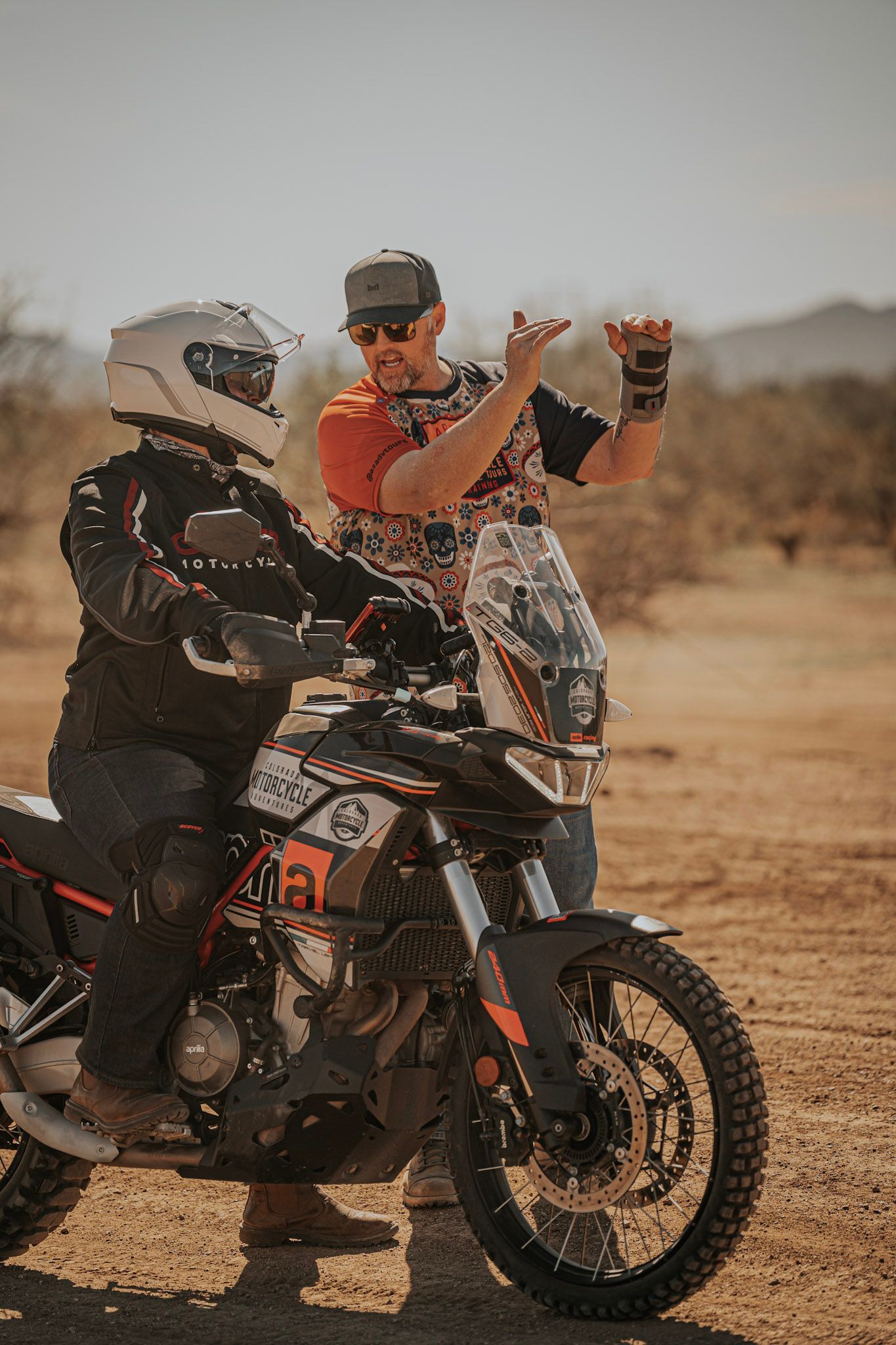 Person on motorcycle, being instructed by another in desert. Motorcycle is black and orange. Bright sunny day.