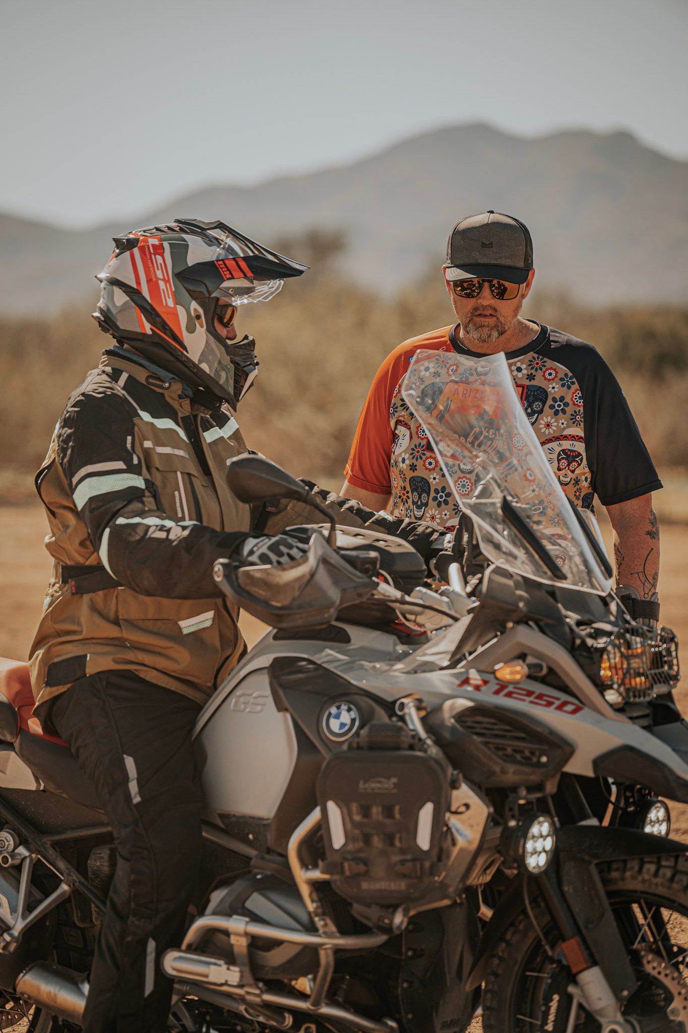 Person in motorcycle gear on a bike, talking to a man in a cap, desert background.