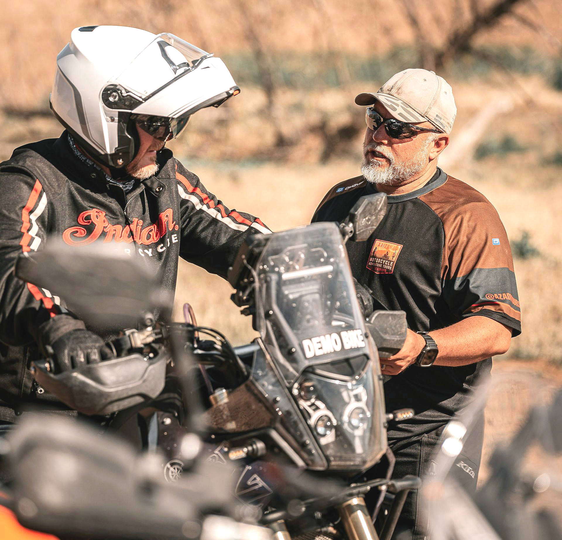 Two men beside a motorcycle, discussing something. One wears a helmet and Indian jacket; the other, a cap and sunglasses.
