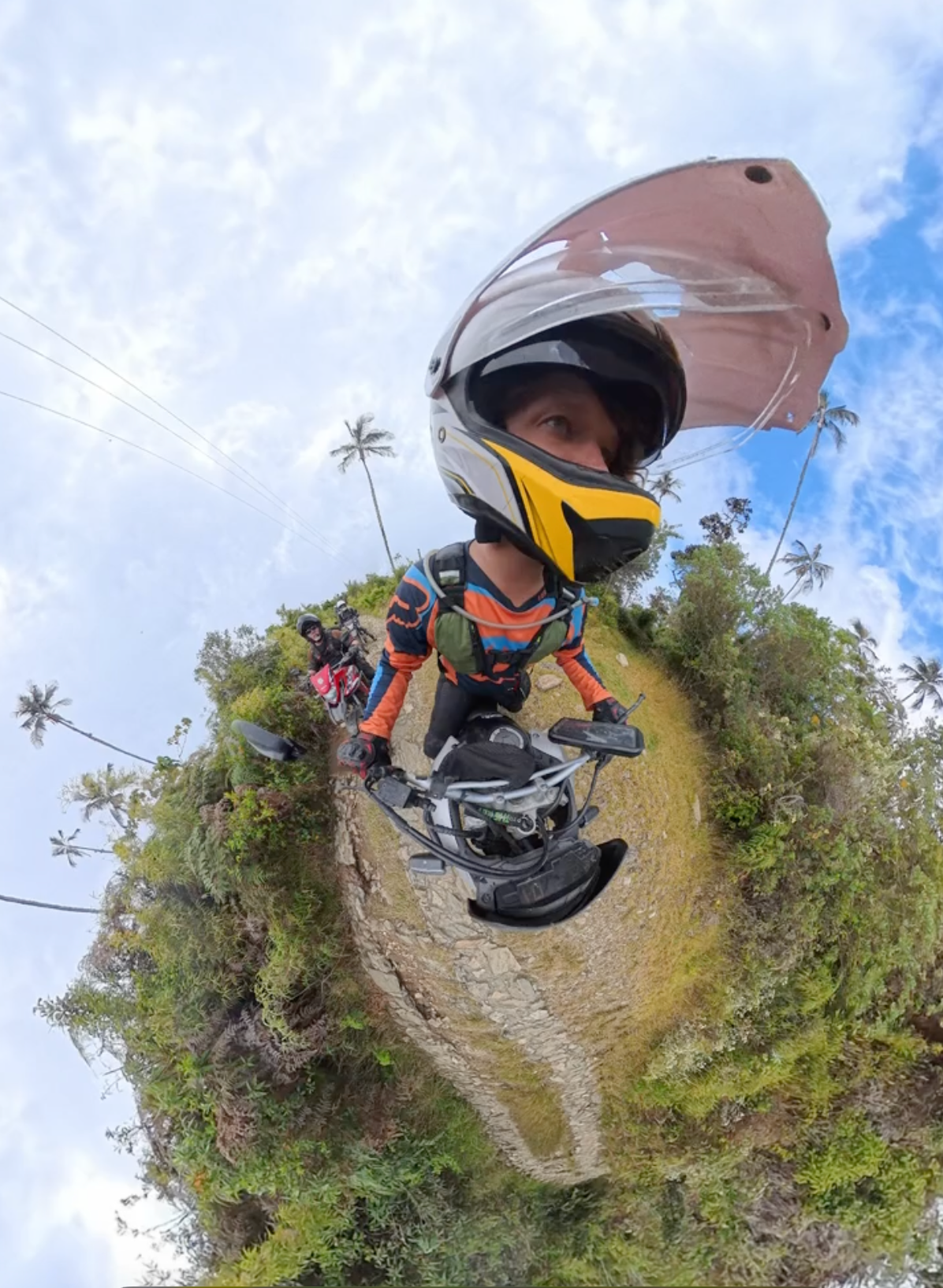 Person on motorcycle, wearing helmet, riding on dirt path in lush green landscape under cloudy sky.