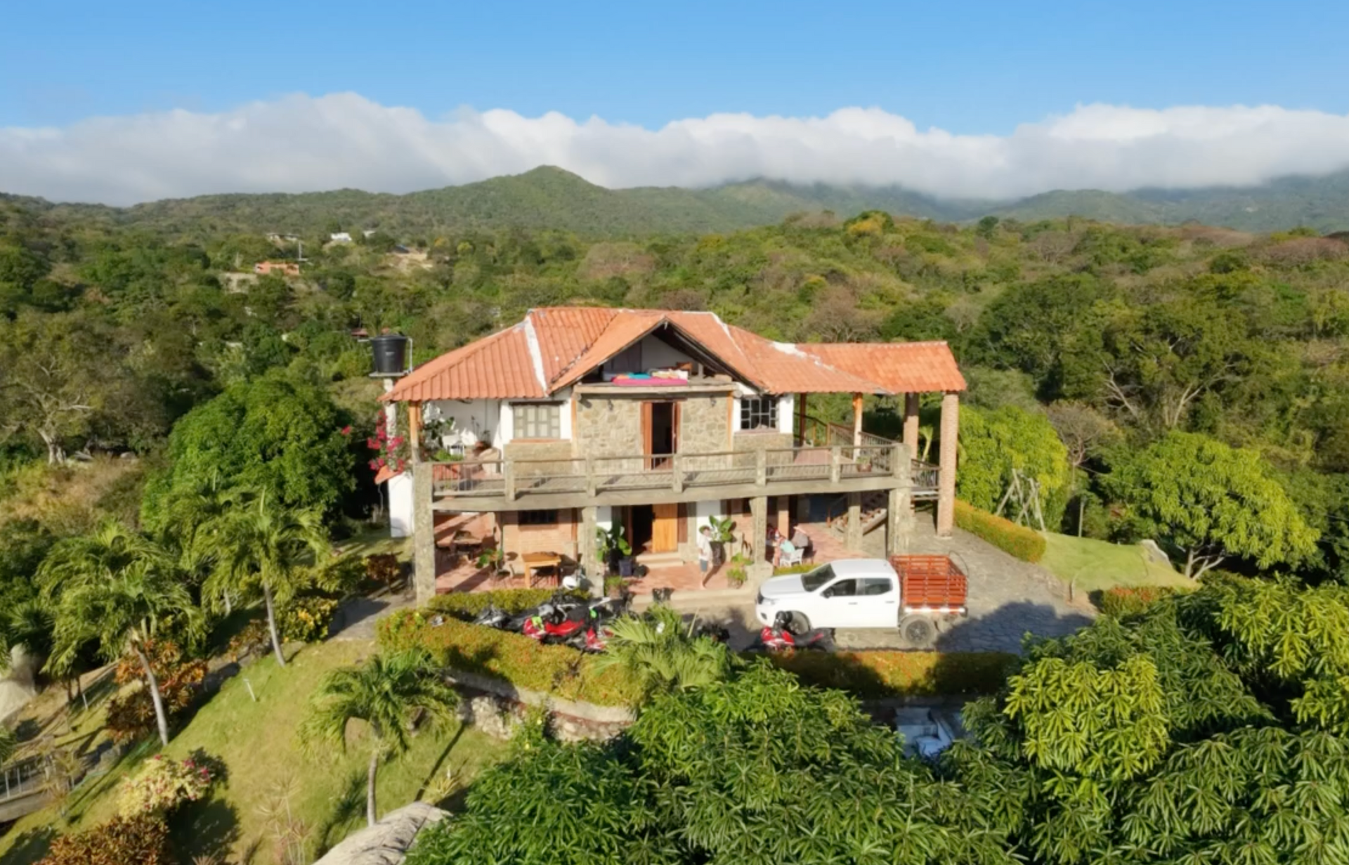 Two-story stone house with tiled roof, balcony, and driveway on a hilltop, surrounded by lush green foliage and mountains.
