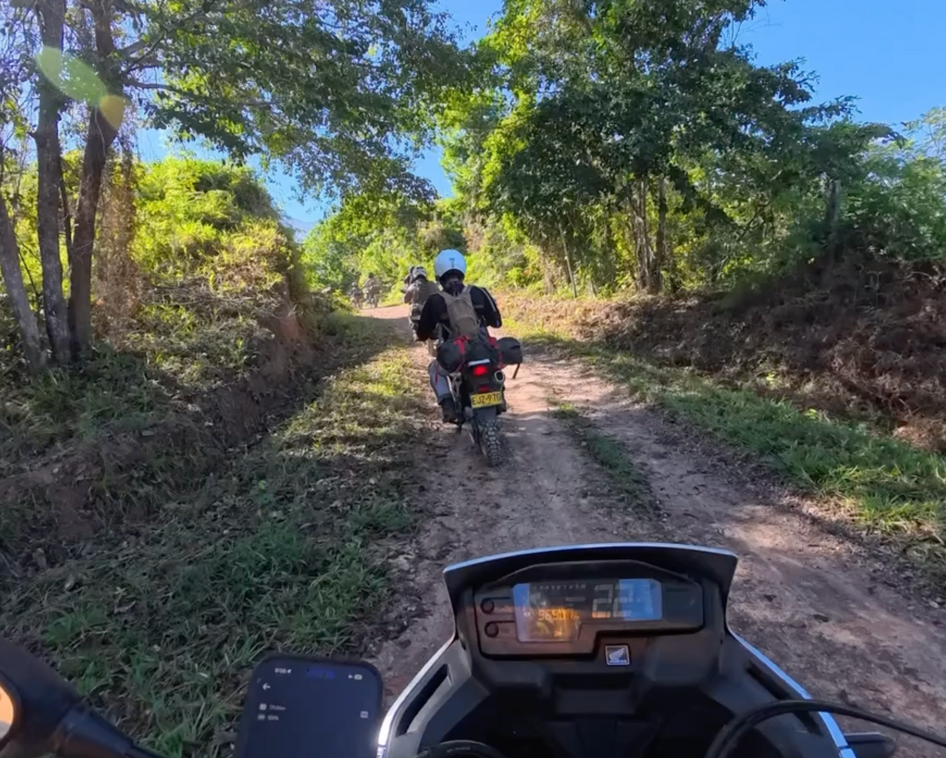 Motorcyclists on a dirt road through a forested area. Sunny day with green foliage and light brown dirt.