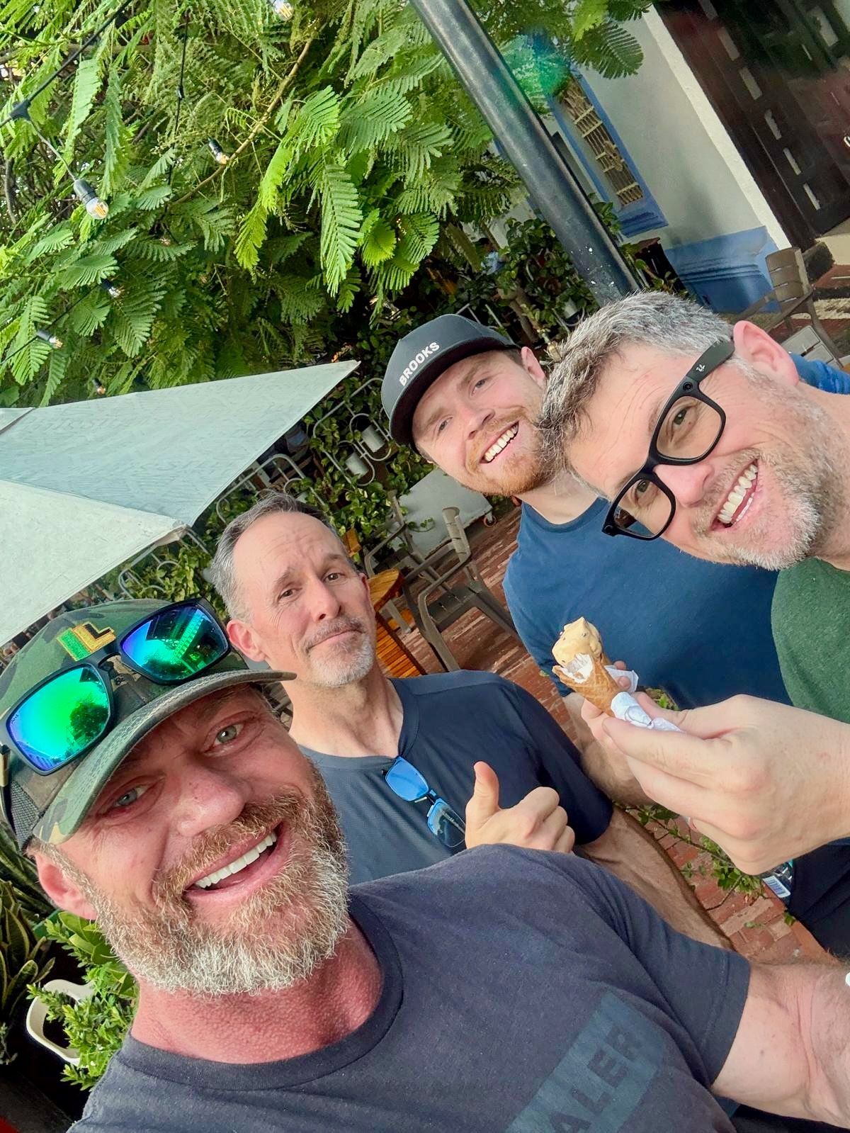 Four smiling men pose for a selfie outdoors. One eats a pastry. Green foliage is in the background.