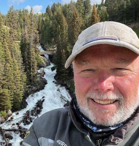 Close up photo of a mans face with a white beard and hat. River and trees in the background.