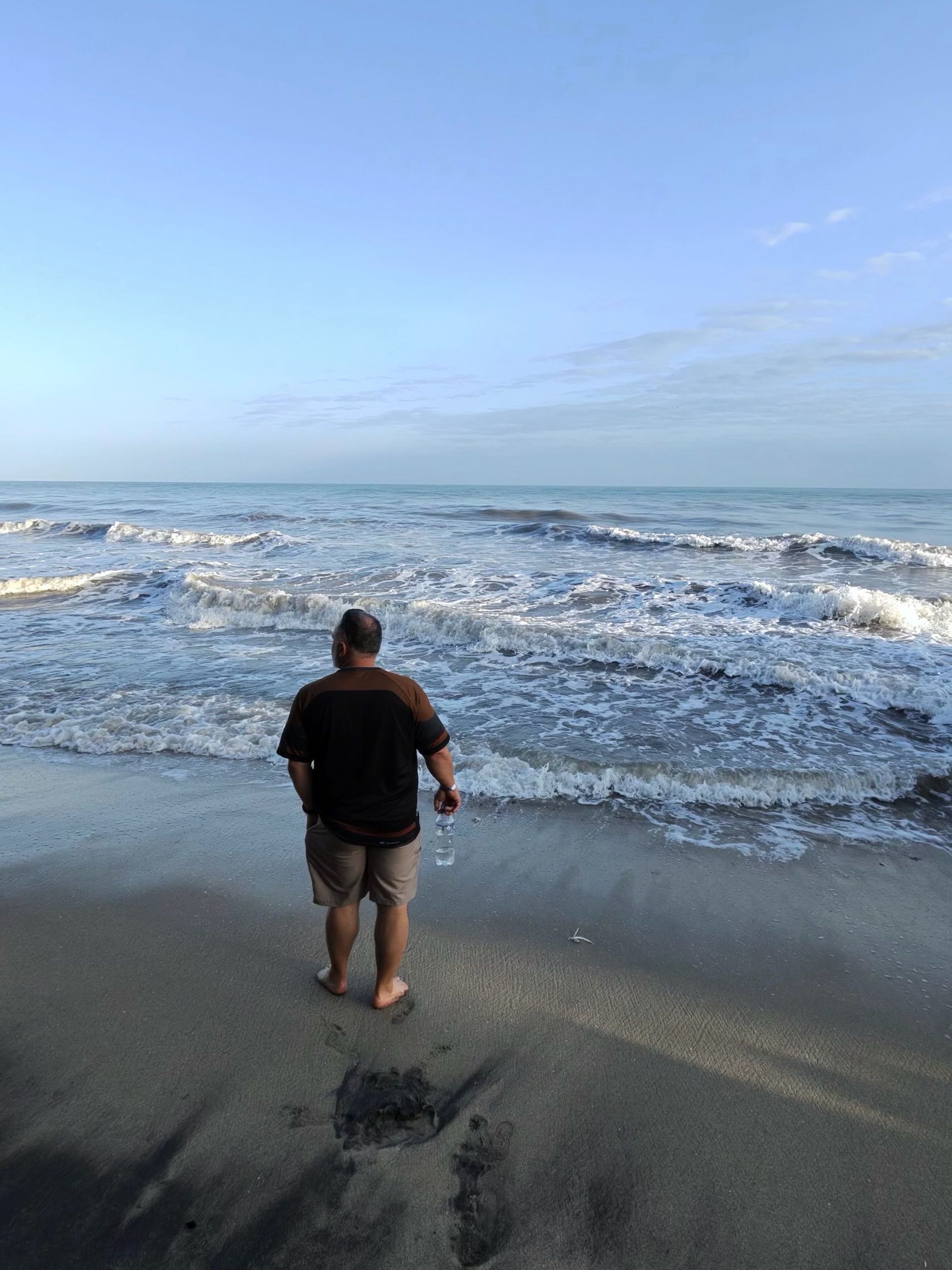 Man standing on beach, looking at ocean. Blue sky, water, and dark sand.