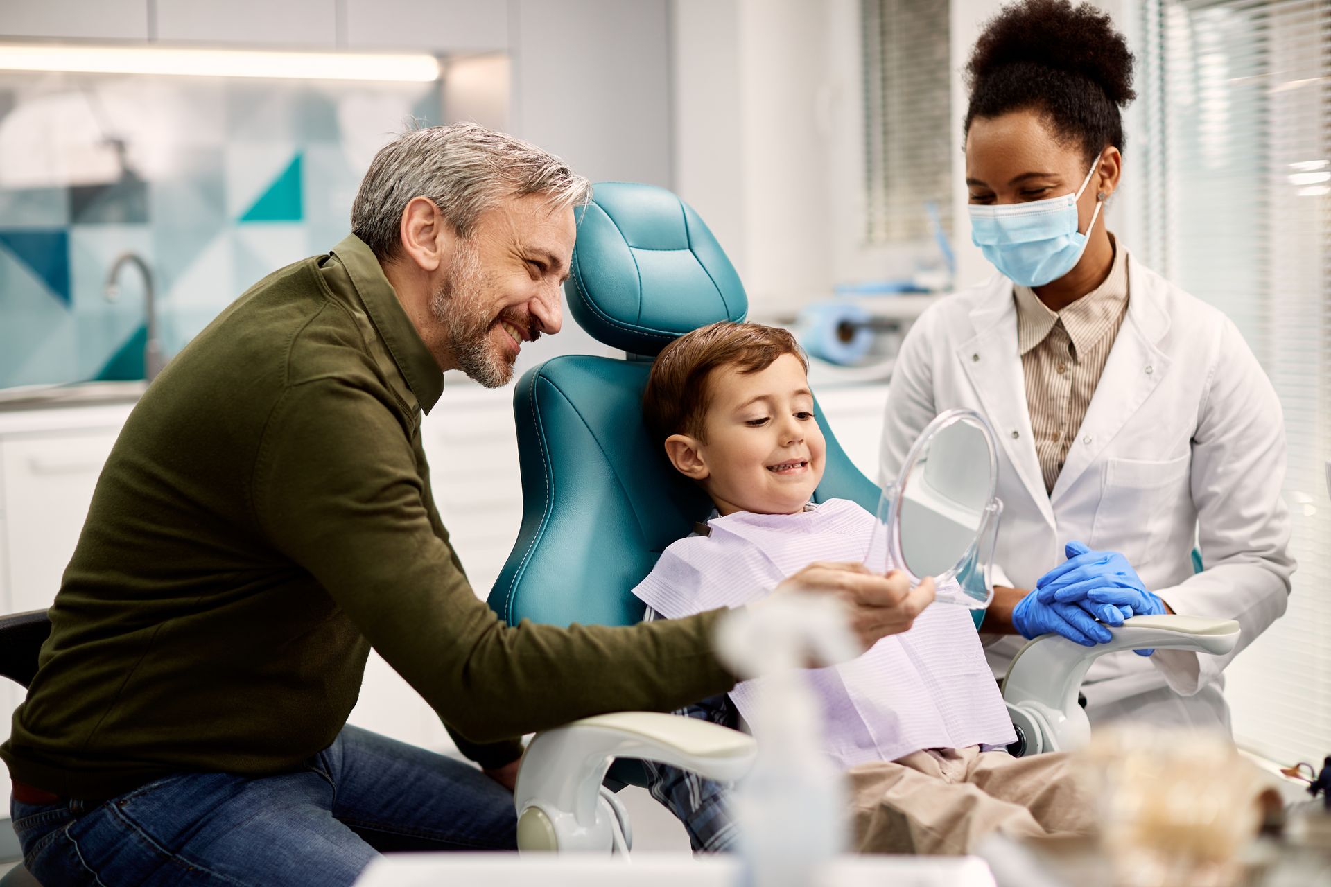 Child seated in a dental chair while a clinician holds a mirror and a caregiver stands nearby.