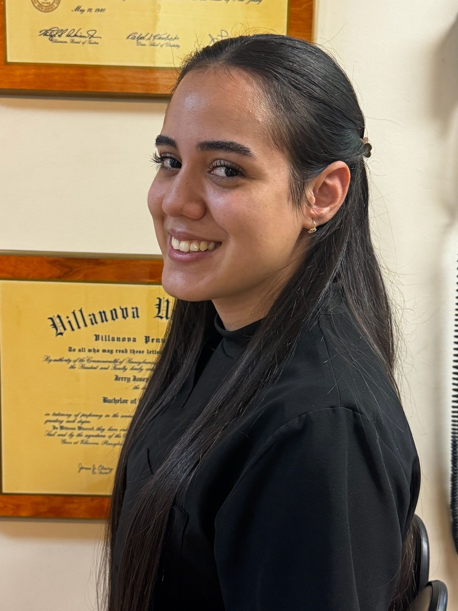 Smiling woman in a black top standing indoors beside framed certificates