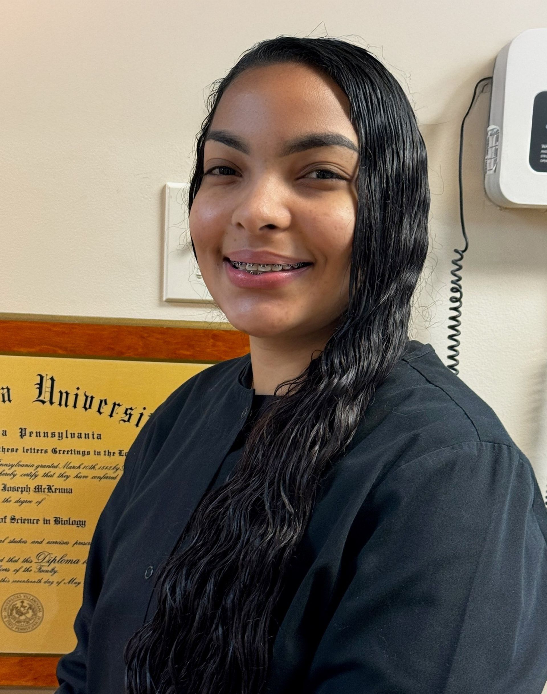 Smiling person in a black top standing indoors beside a framed certificate on the wall