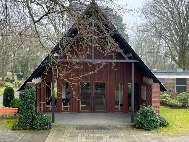 Friedhofskapelle: ein Holzhaus mit spitzem Giebeldach, Gastüren und rotem Backstein, umgeben von Bäumen und Sträuchern.