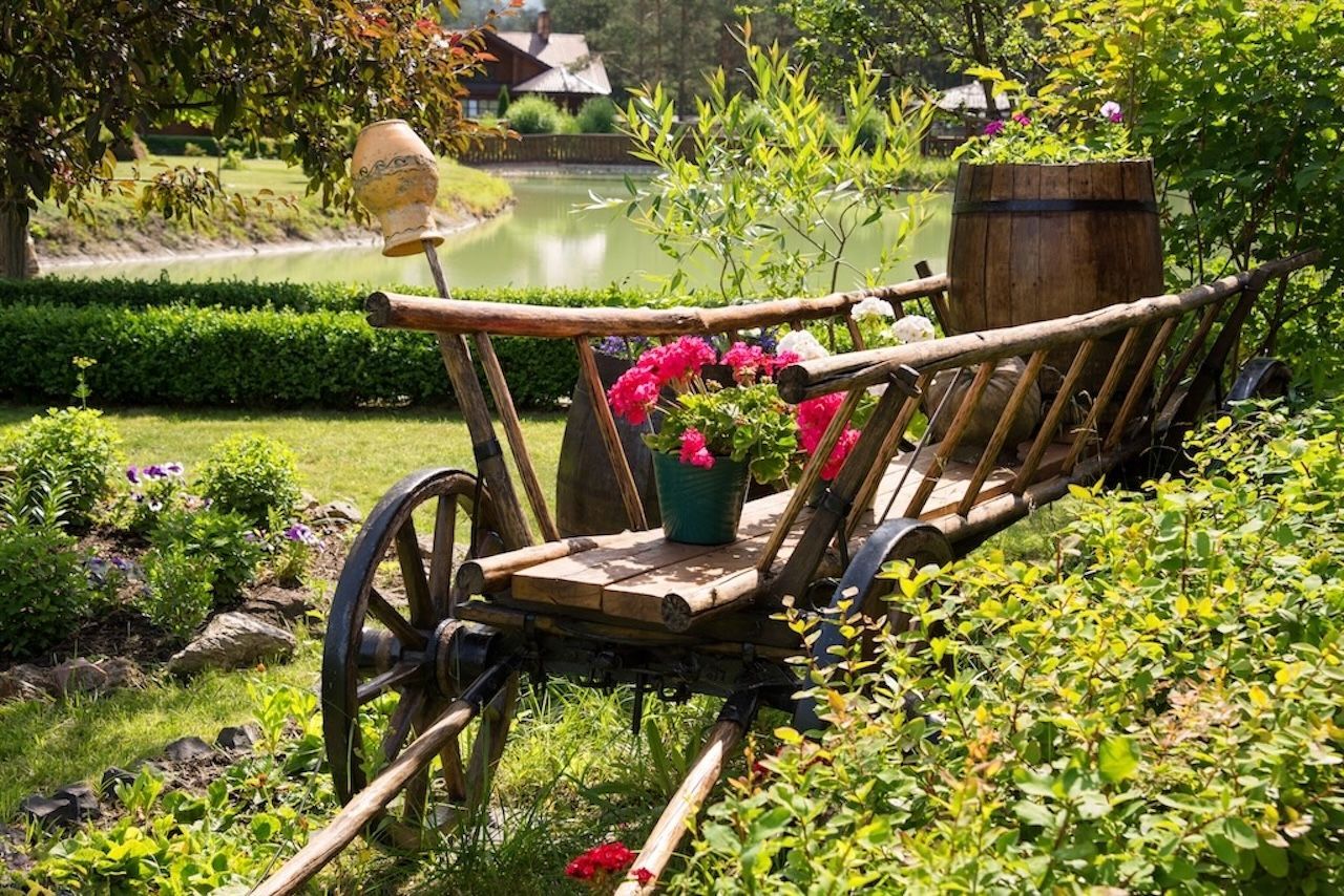 Alter hölzerner Heuwagen mit großen Speichenrädern. Bepflanzt mit roten Blumen und mit einem Holzfass beladen im Garten.