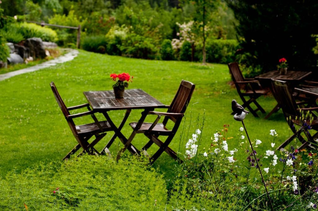 Tischgruppe mit  Stühlen auf grünem Rasen, dekoriert mit einem Blumentopf mit roten Blumen umgeben von Blumen und Sträuchern.