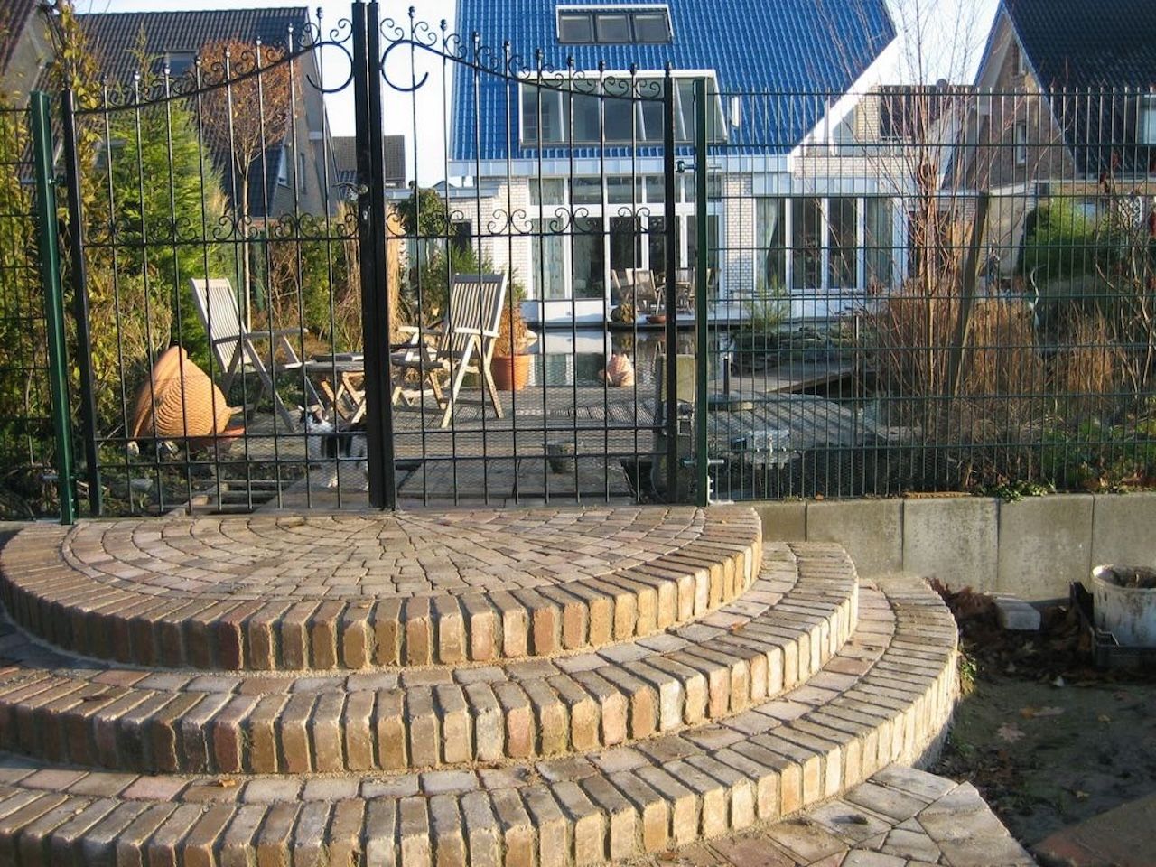 Halbrunde Treppe aus Naturstein vor schwarzen Metallzaun mit geschwungenen Ornamenten. Dahinter ein Haus mit blauem Dach.