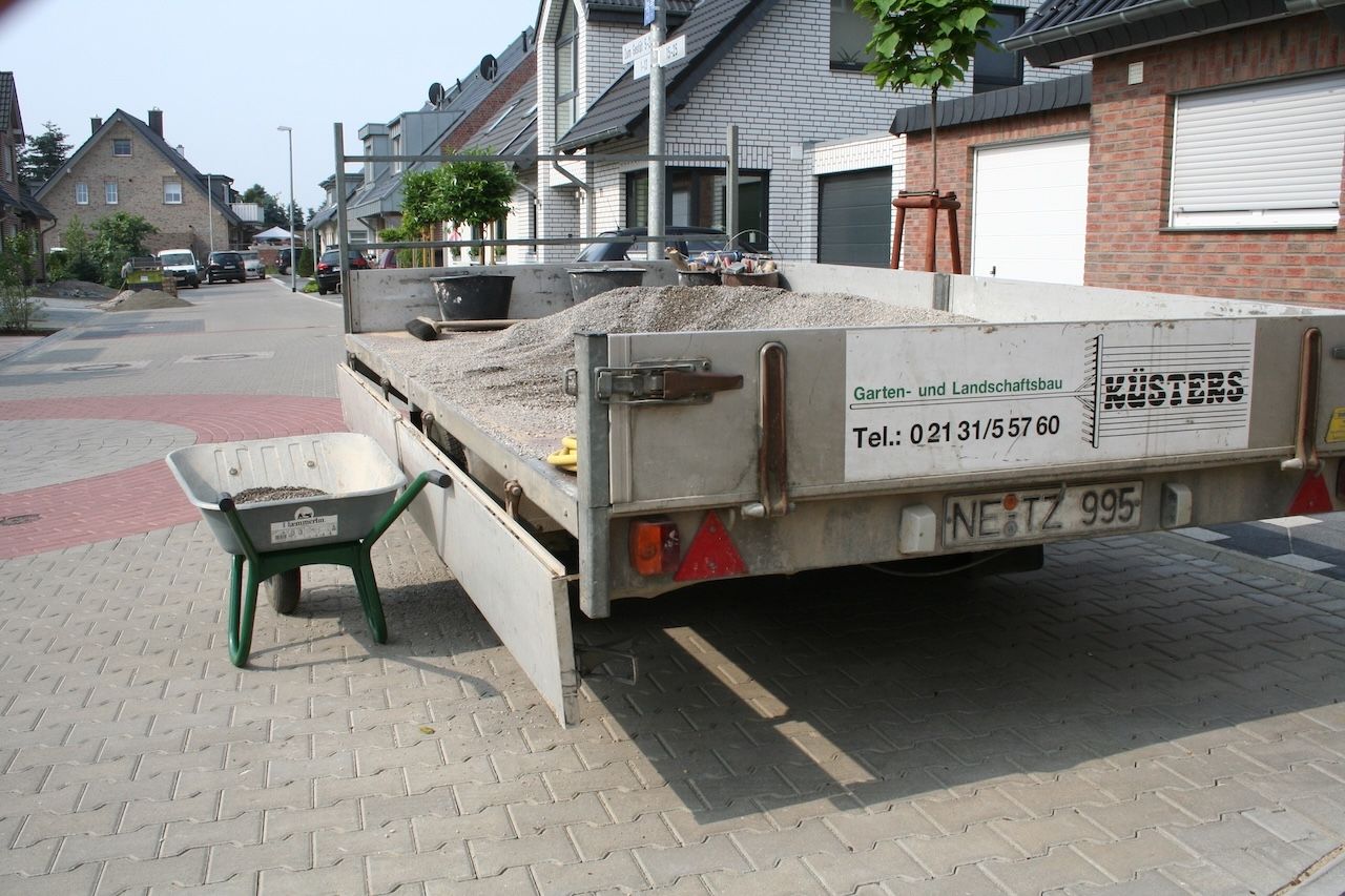 Offener Anhänger mit Kiesladung und Aufschrift 'KÜSTERS Garten- und Landschaftsbau' auf gepflasterter Straße mit Schubkarre.