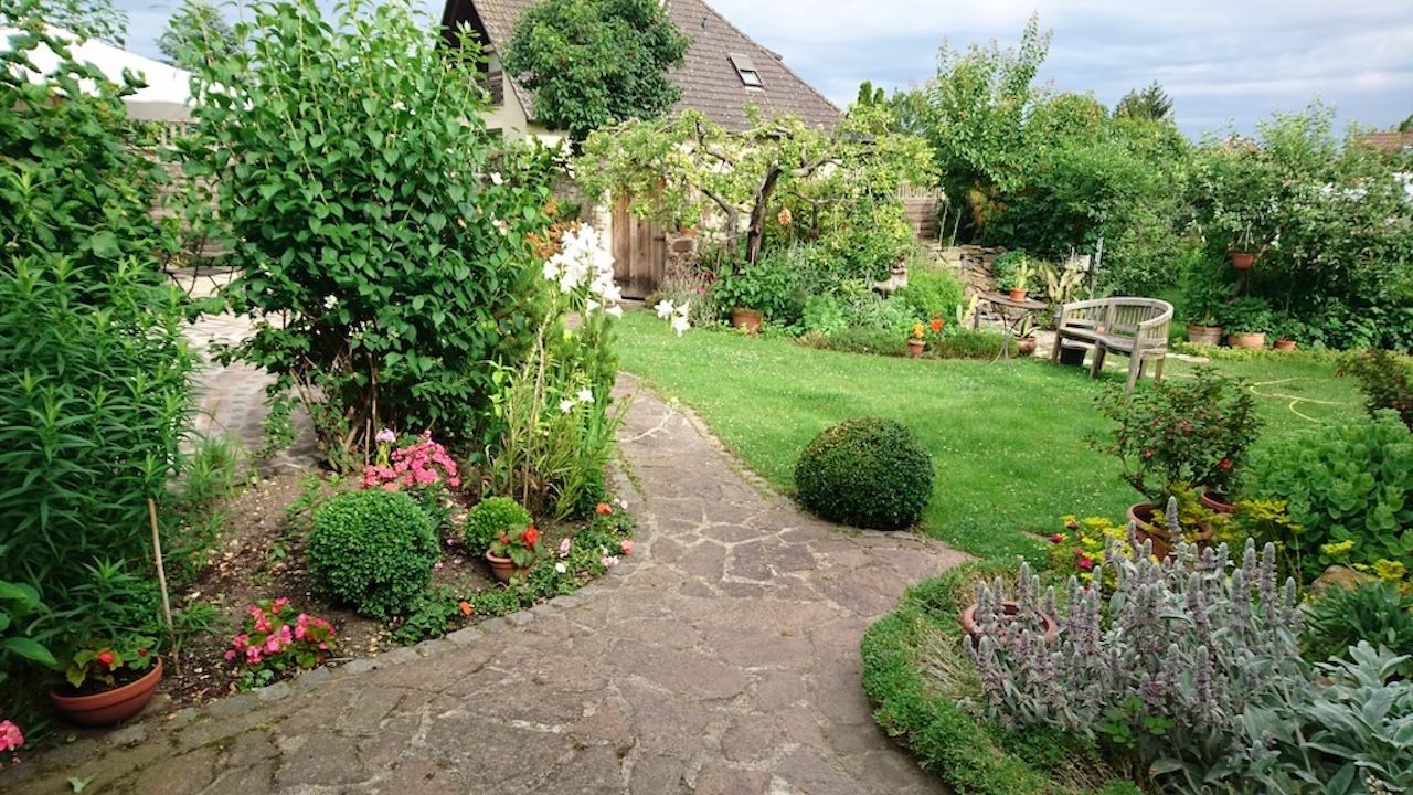 Garten mit geschwungenem Natursteinweg, umgeben von grünen Sträuchern, Blumen und Rasenflächen, im Hintergrund Gartenmöbel.