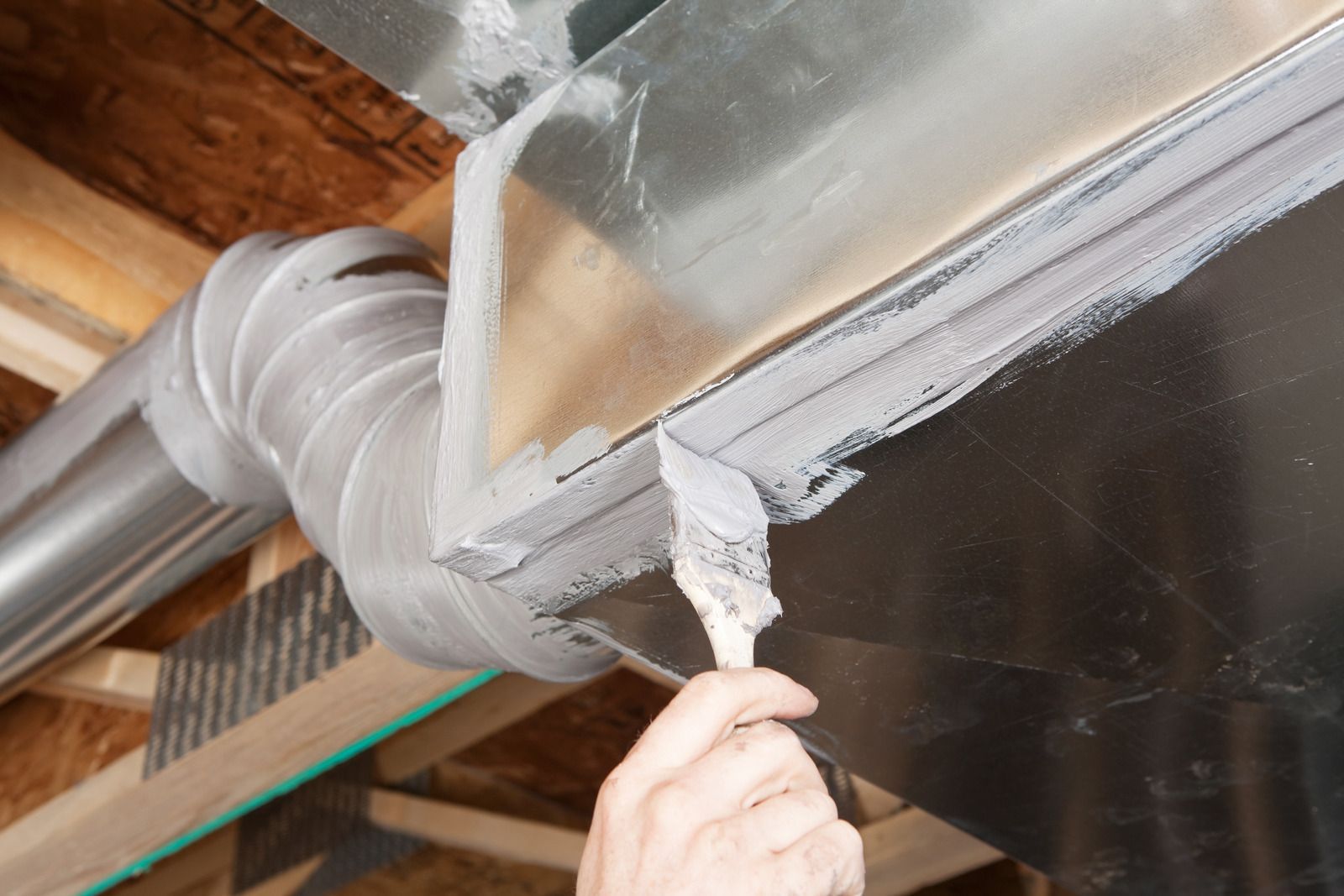 Person applying gray sealant to metal ductwork in an unfinished attic space.