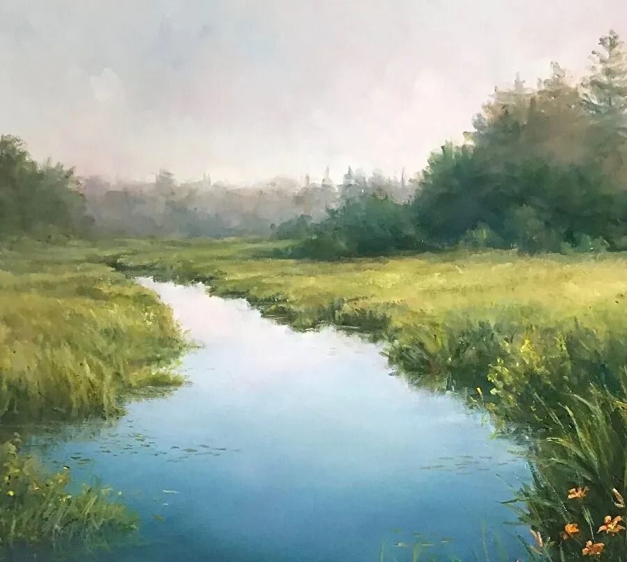 Calm stream flows through a grassy meadow; trees in the background under a hazy sky.