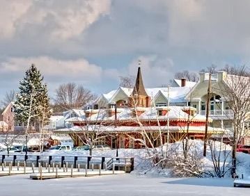 Snowy scene of a lakeside building with a steeple, trees, and a pier.