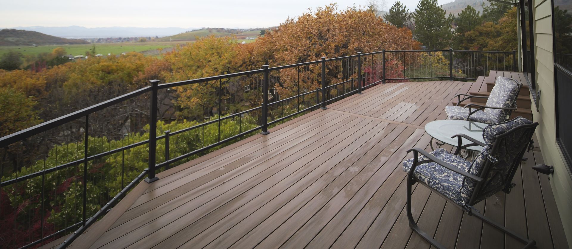 A wooden deck with black railing and chairs overlooks a green landscape with fall foliage.