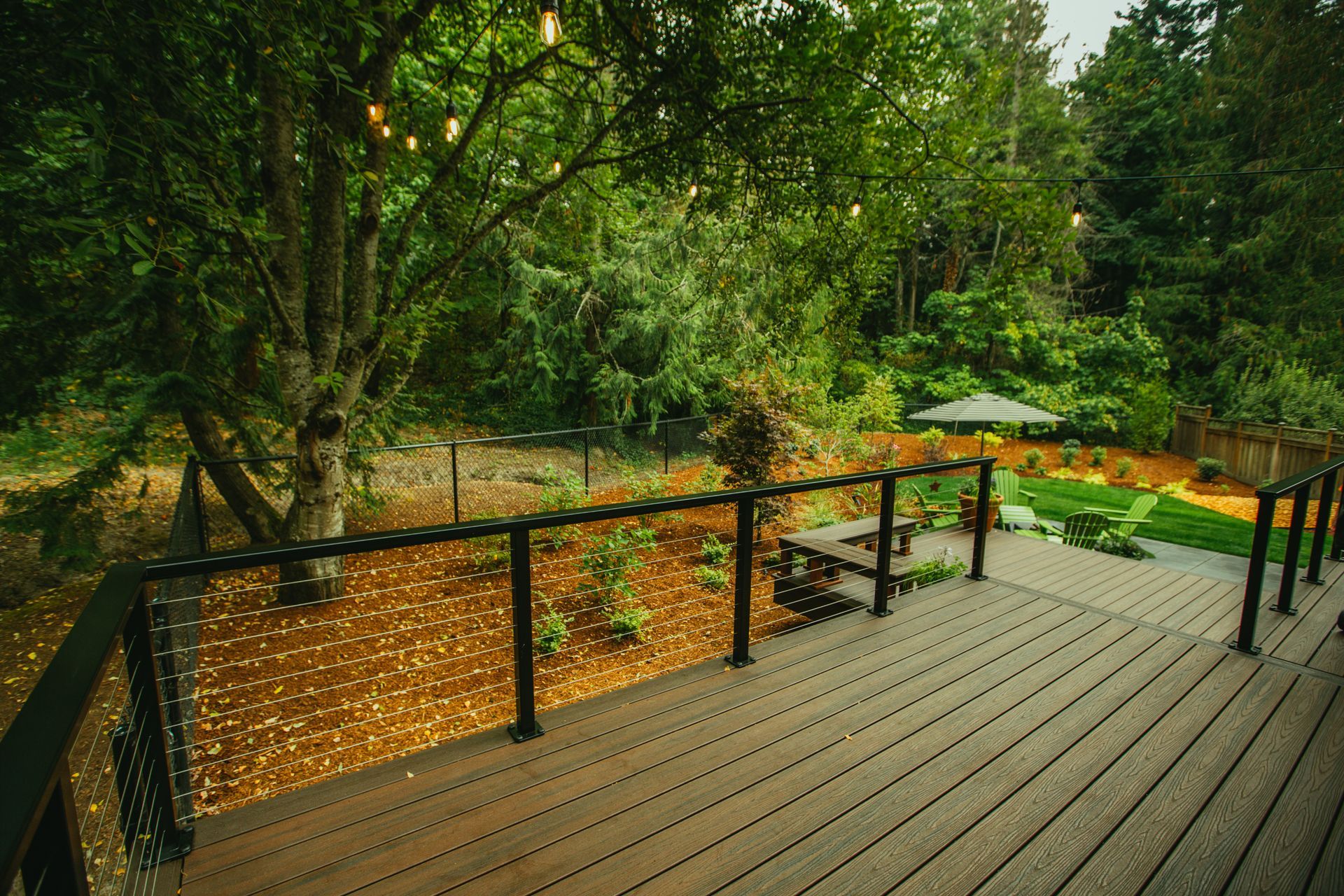 Wooden deck overlooking a lush backyard with trees, grass, and a small patio with an umbrella.