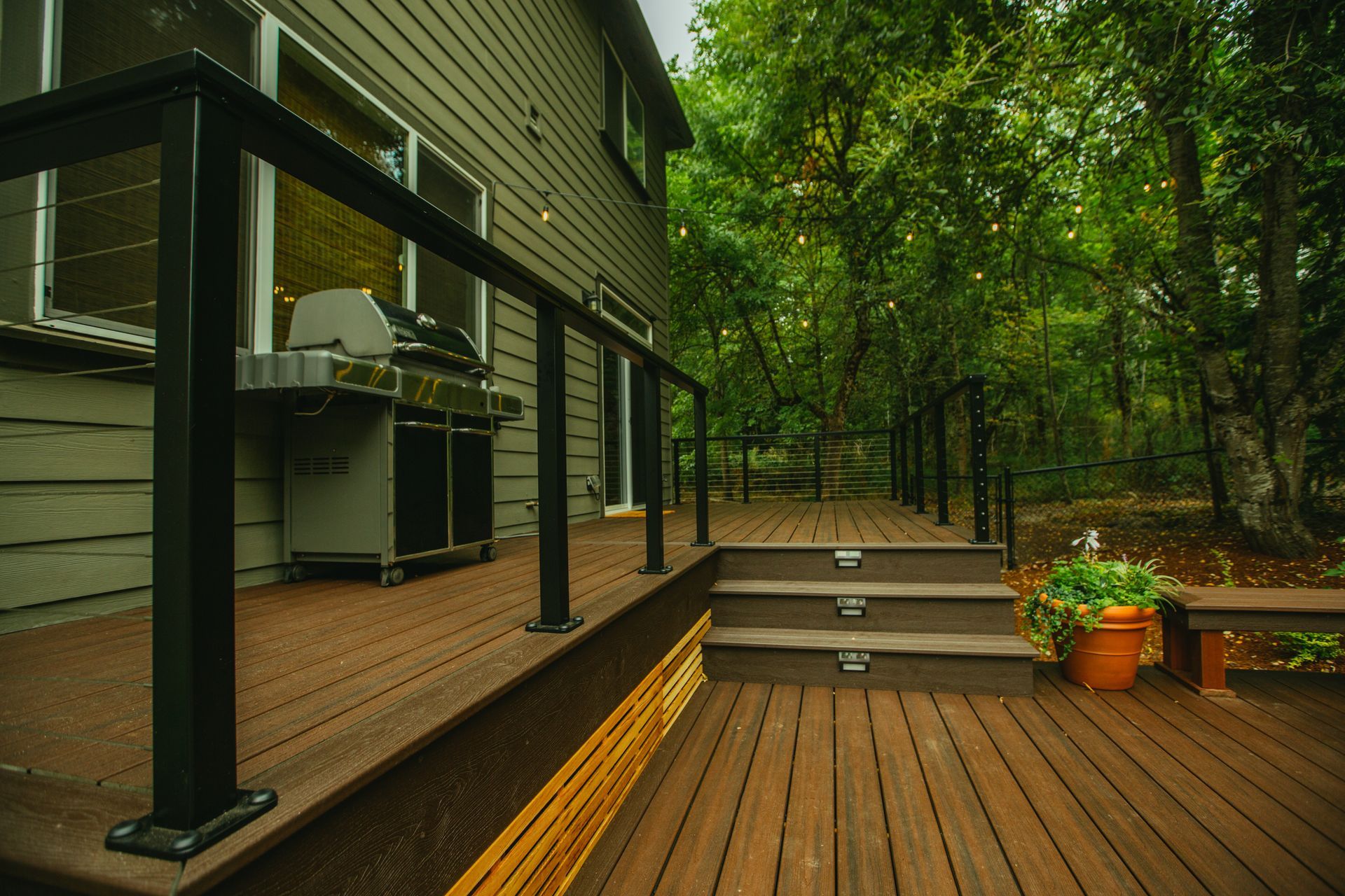 Brown deck with black railing, steps leading to a wooded area; a grill sits next to the house.