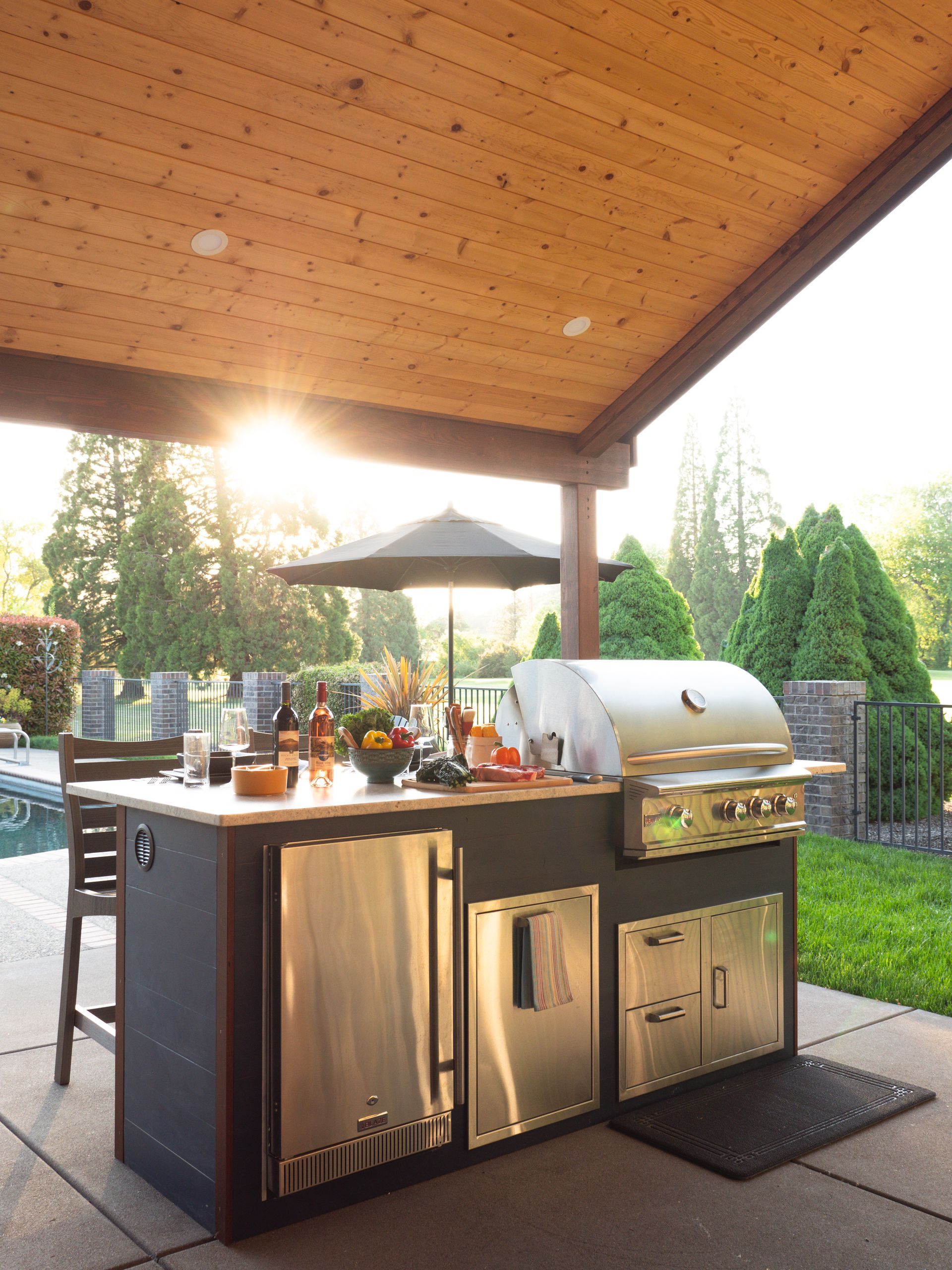 Outdoor kitchen with grill, refrigerator, and prep space under a wood-paneled awning; backyard setting.