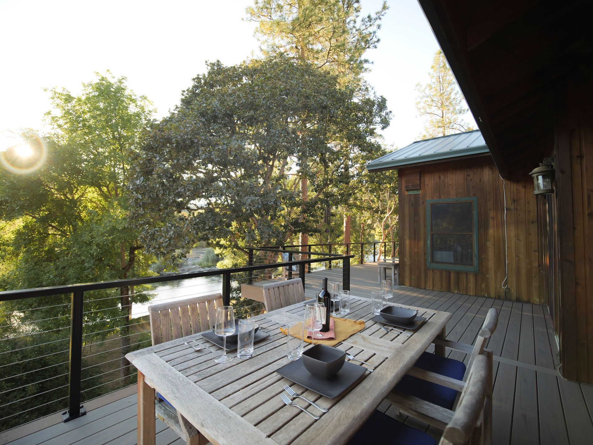 Wooden deck with dining table set for a meal overlooking a river and trees, sunlight.