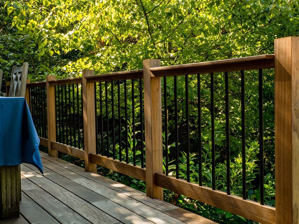 Wooden deck railing with black vertical bars, overlooking lush green foliage.