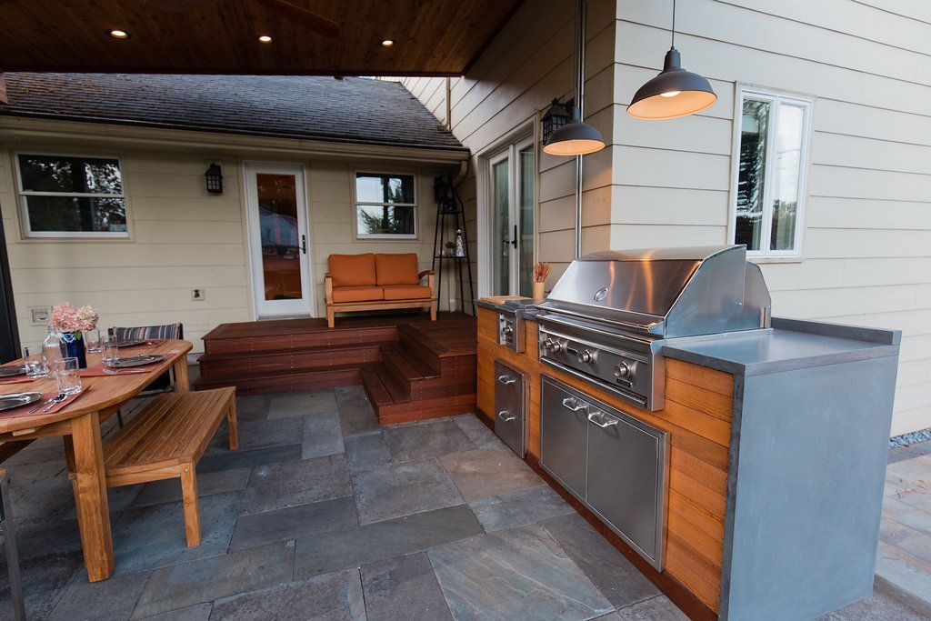 Outdoor kitchen with grill, wooden cabinetry, dining table, and bench on a stone patio.