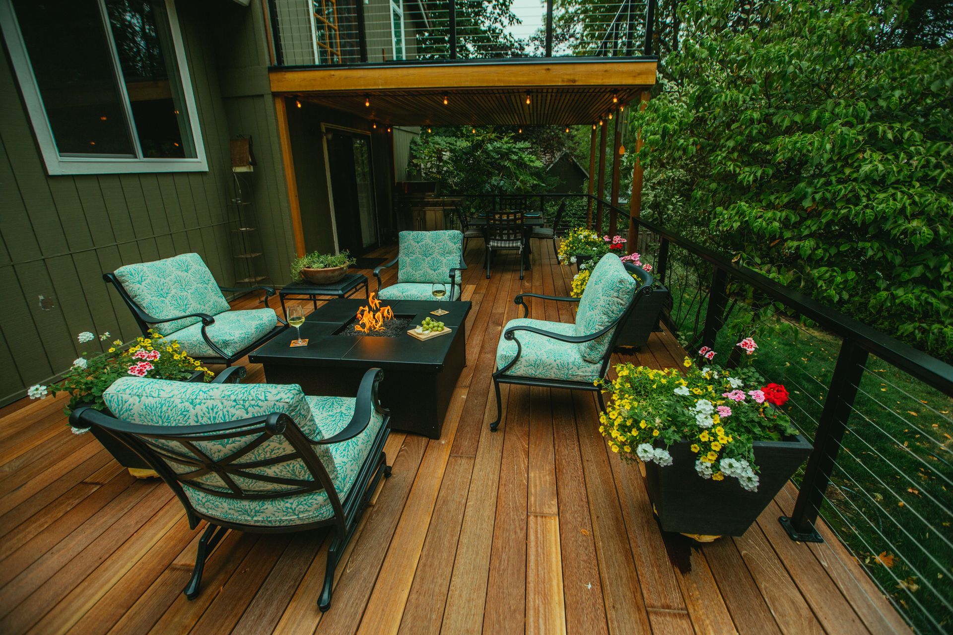 Wooden deck with seating area, fire pit, and potted flowers. Green chairs, natural wood, and lush greenery.