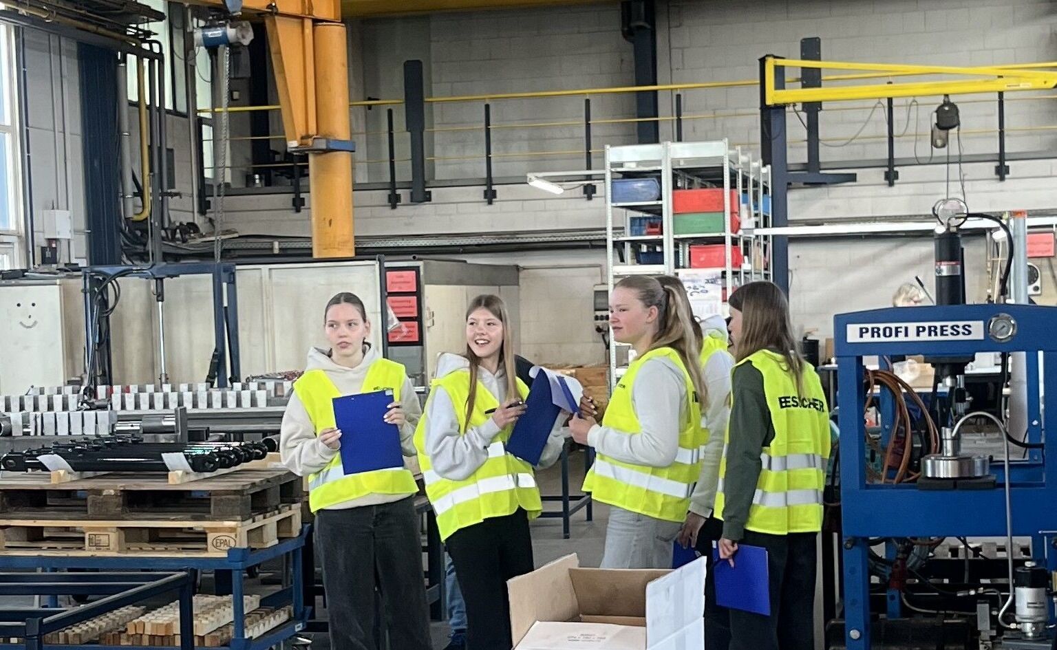 Vier Personen in Warnwesten stehen in einer Industriehalle und unterhalten sich in der Nähe von Maschinen.