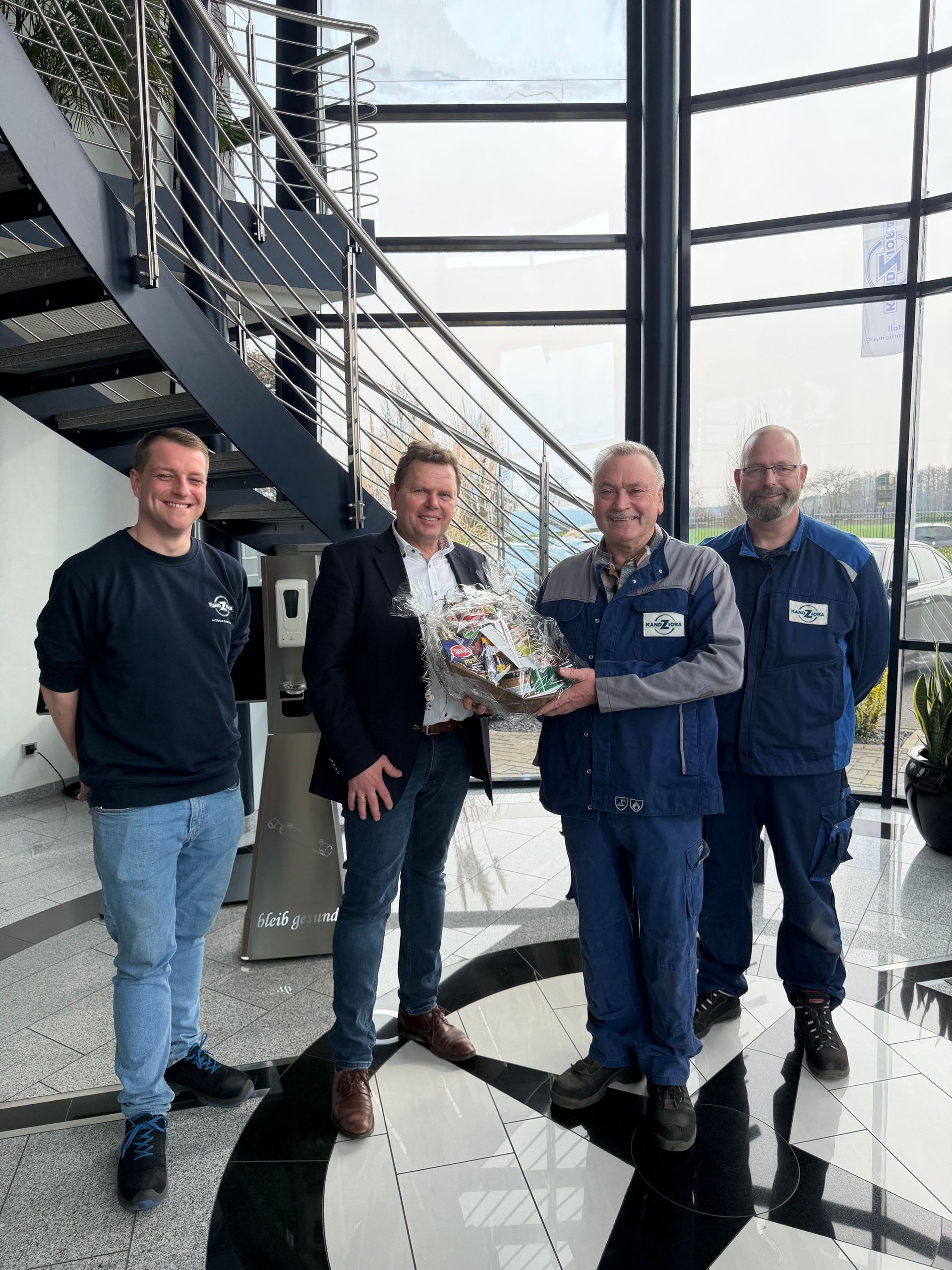 Vier Personen stehen in einem hellen, modernen Büro mit einer Treppe, eine hält einen Geschenkkorb für eine andere.