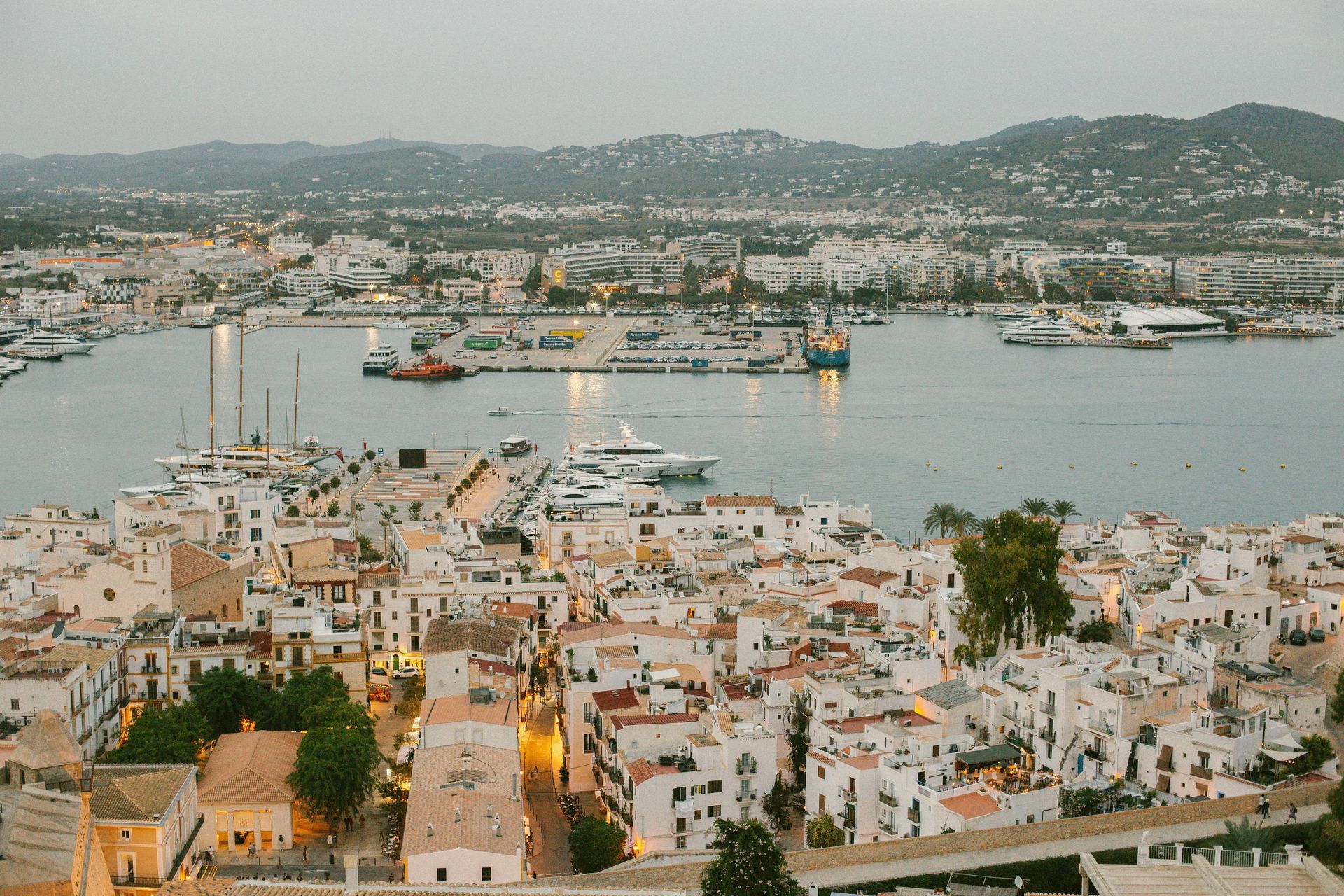 Panoramisch uitzicht op Ibiza-stad, met witte gebouwen, boten in de haven en heuvels in de verte.