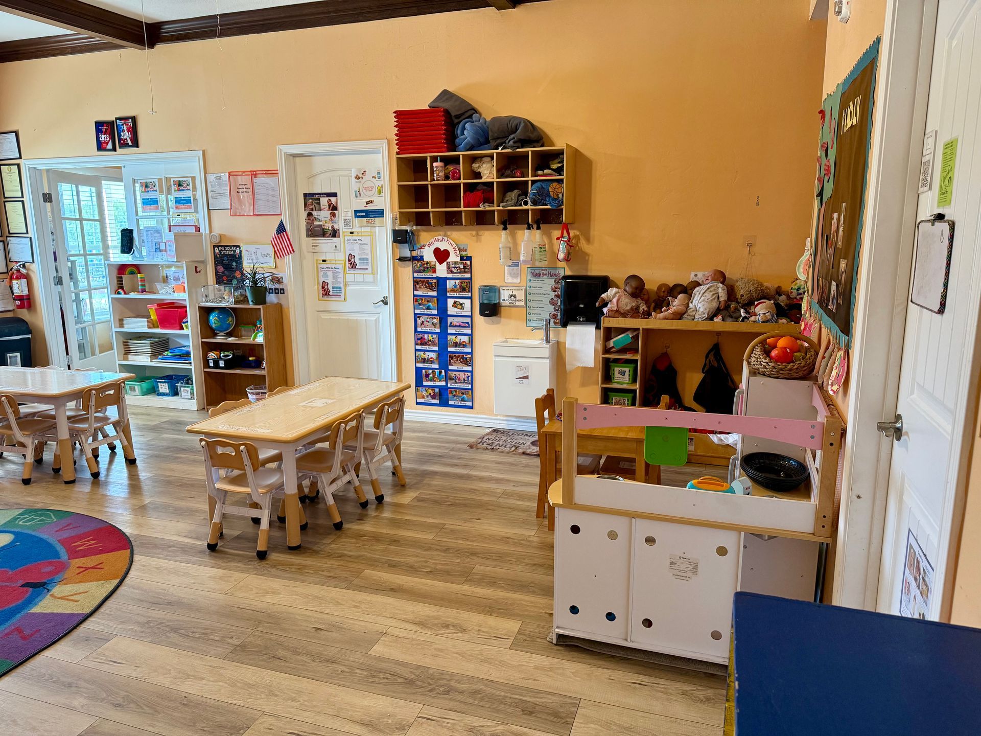 A colorful preschool classroom with small tables, cabinets, and shelves filled with toys and art.
