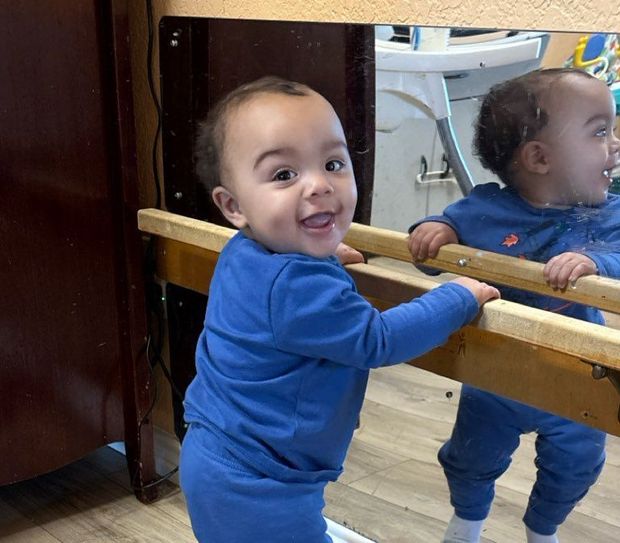 A baby is standing in front of a mirror and smiling
