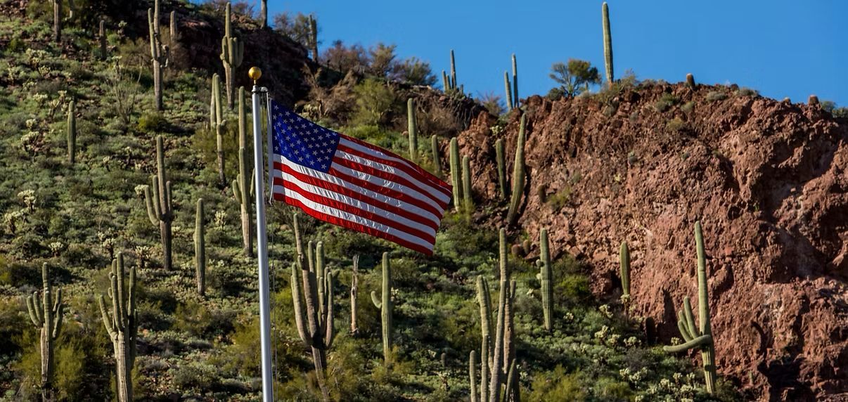 American flag on a pole amid desert cacti and rocky hills under a clear blue sky