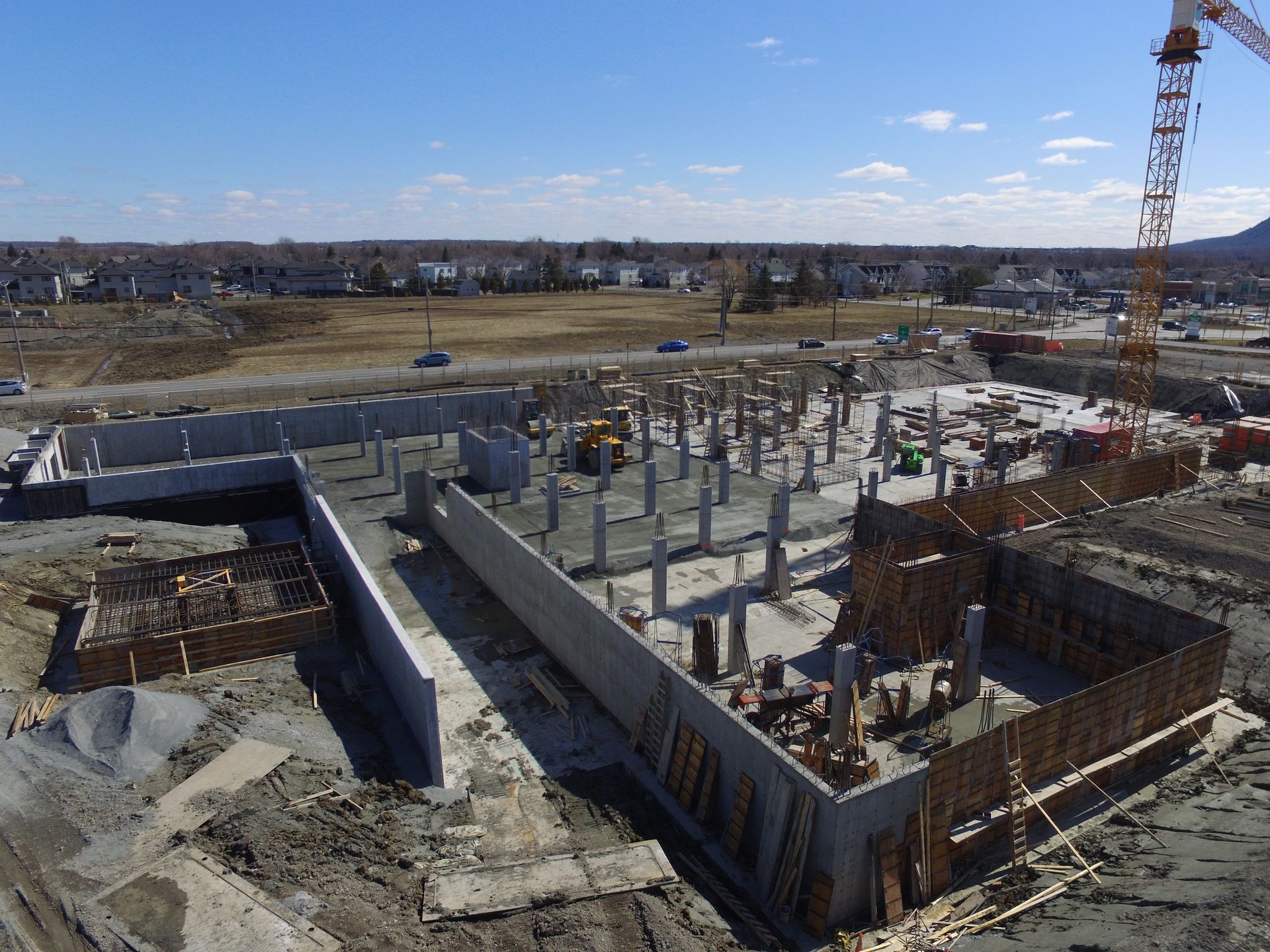 Chantier de construction avec murs de fondation, colonnes en béton et grue, sous un ciel bleu clair.