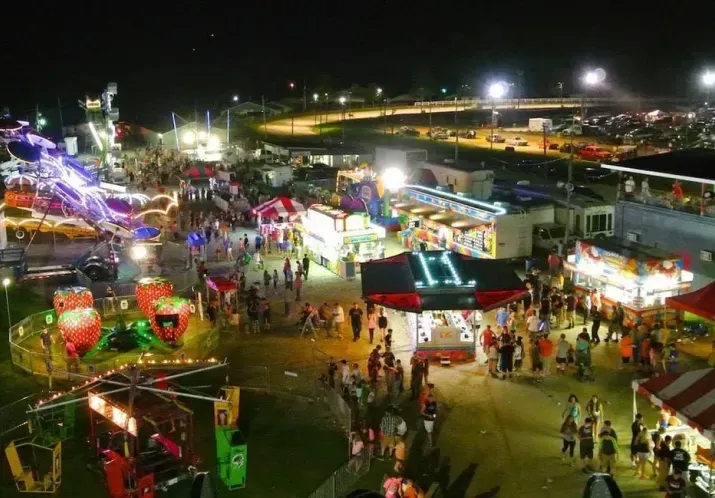Nighttime fairground with bright rides, colorful booths, and crowds walking among attractions.