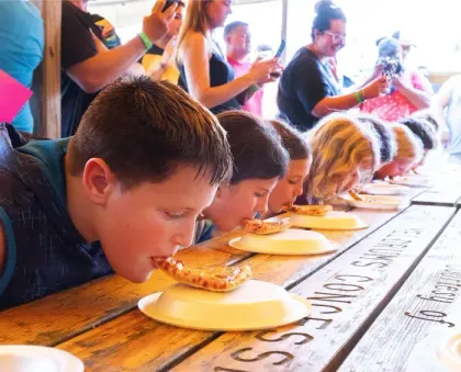 Children leaning over plates, eating noodles at a long wooden table in a bright dining hall.