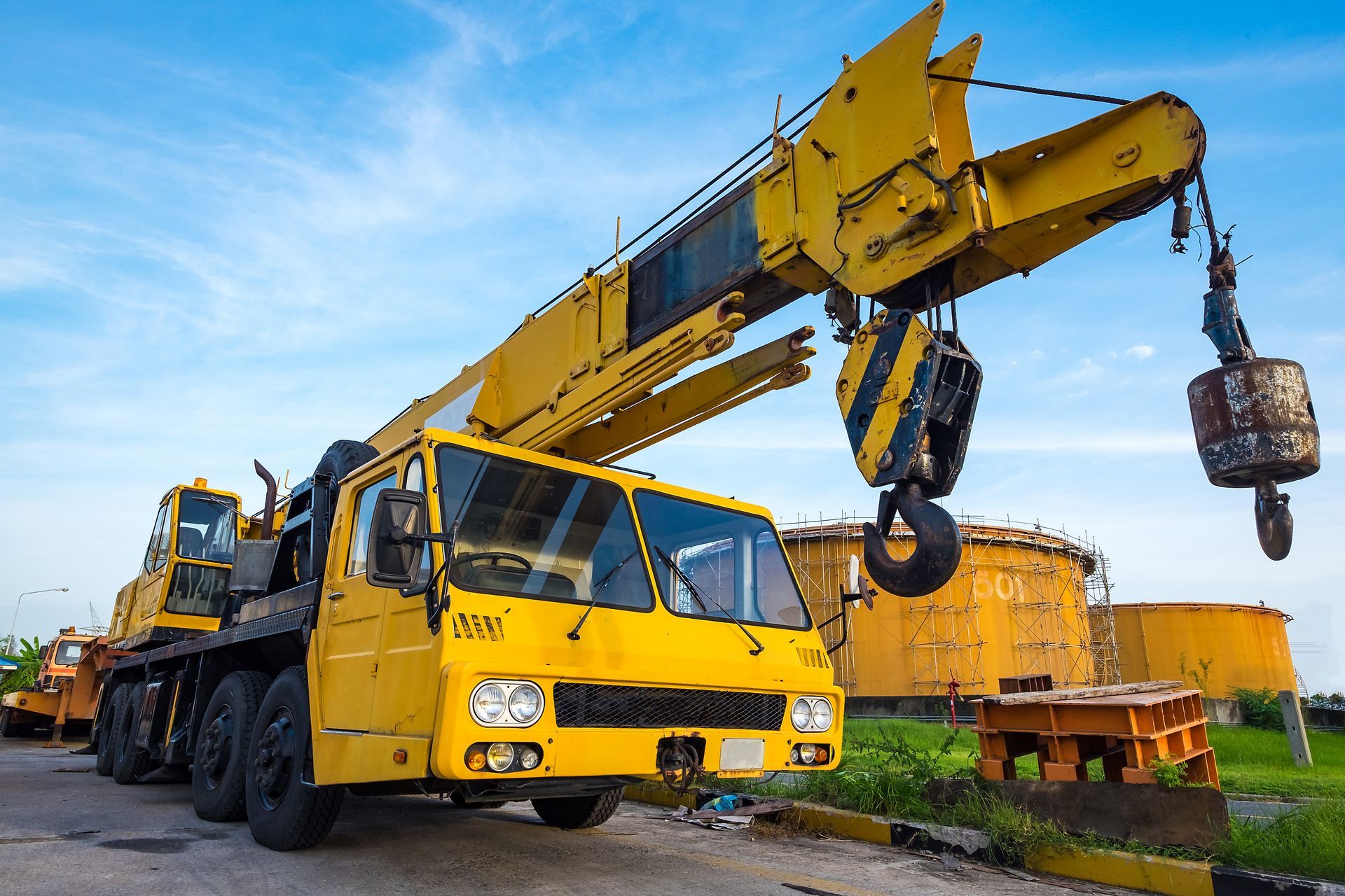 A yellow mobile crane is on a lot with grass and fuel tanks, against the blue sky.