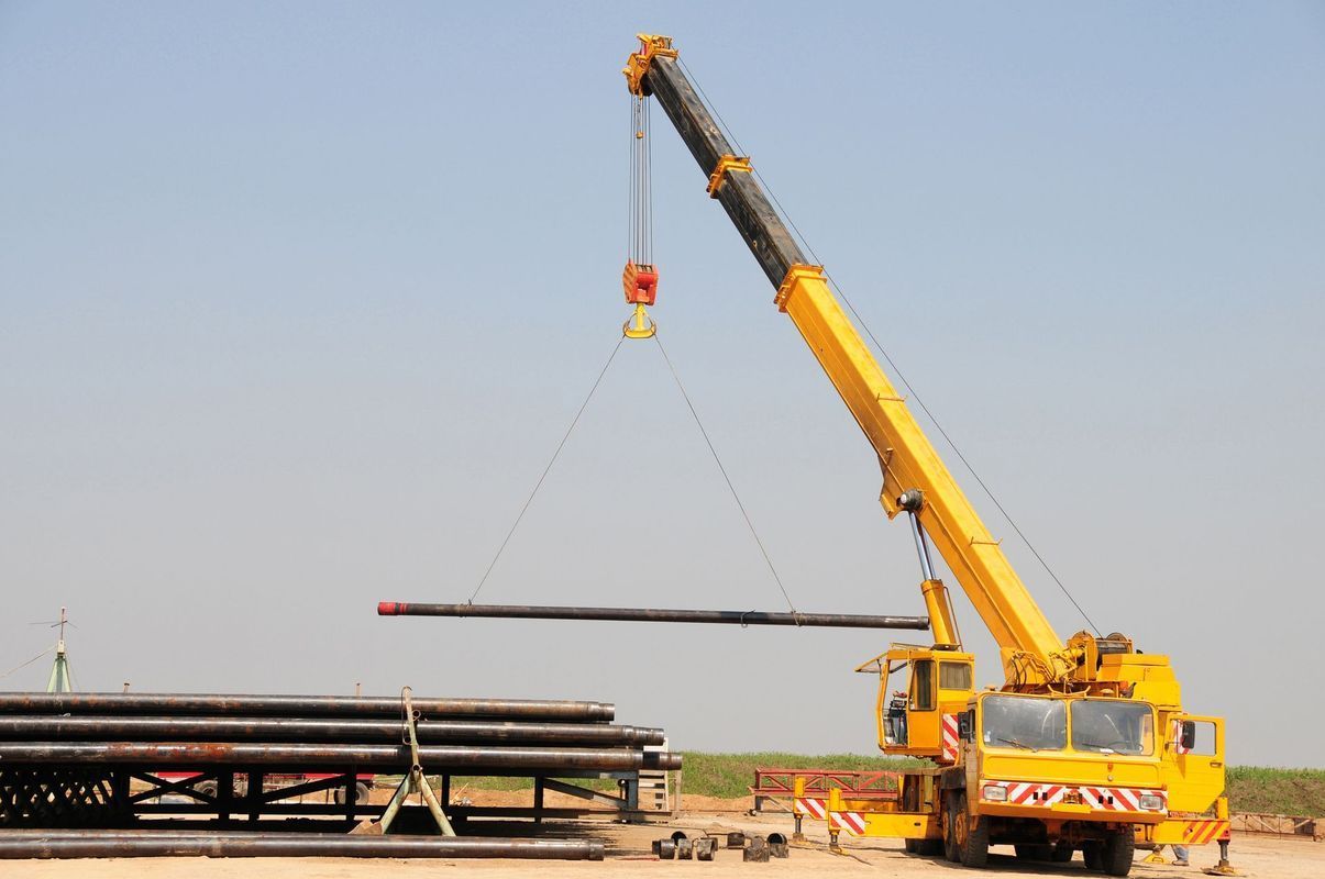 Yellow crane lifting a long metal pipe from a pile outdoors.