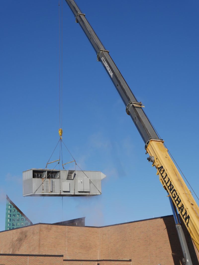 A crane lifting a large HVAC unit above a building on a clear, blue sky day.